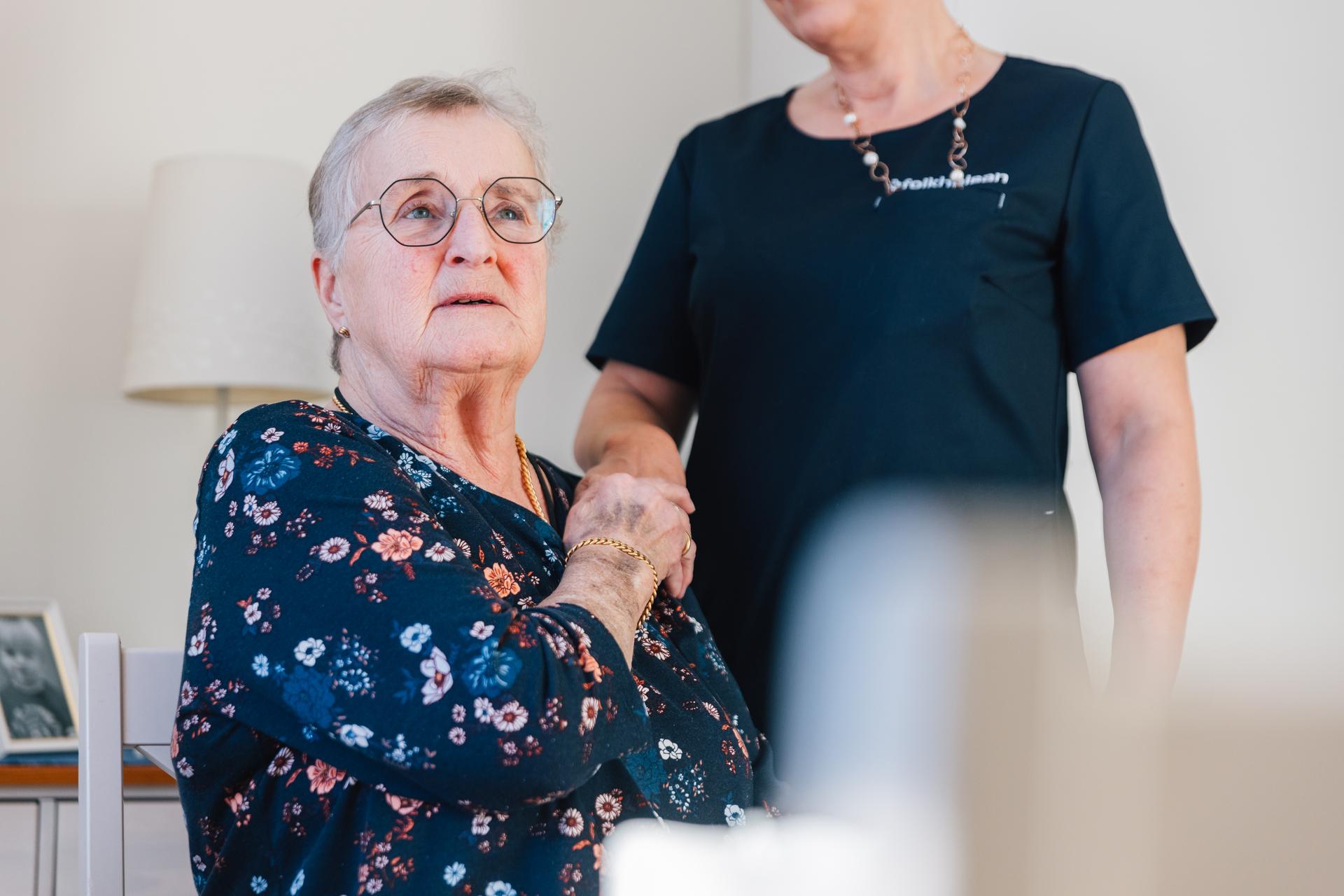 En äldre person sitter vid ett bord och håller händerna samman. Bredvid står en person i mörk arbetsklädsel med Folkhälsan-logotyp, med ena handen lätt vilande på den sittandes axel. Båda bär glasögon. I bakgrunden syns ett vitt skåp och några inramade fotografier.