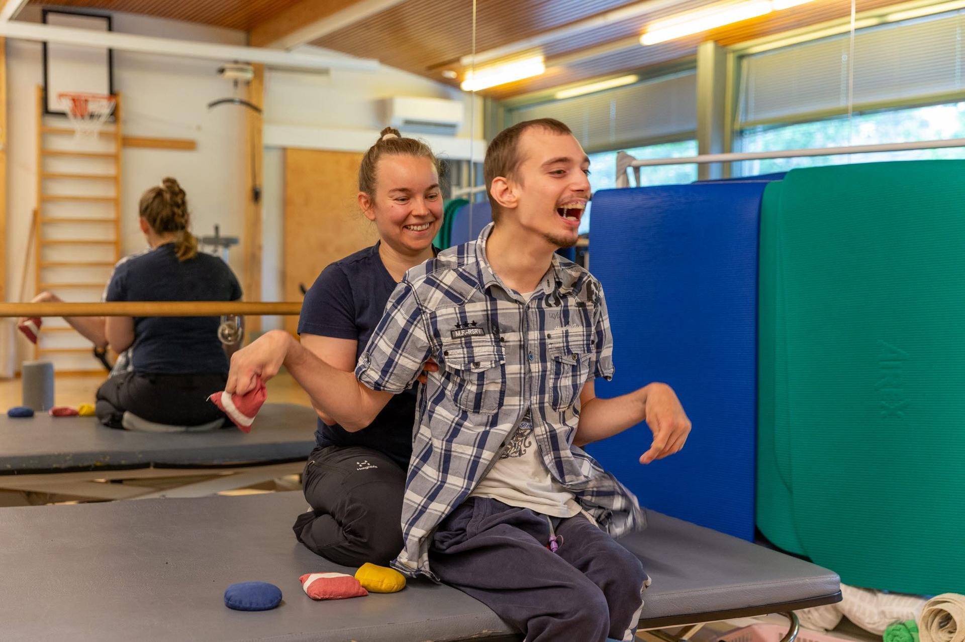 En person sitter på en brits med stöd av en terapeut bakom sig. Små färgade viktsäckar ligger på britsen i en träningssal.