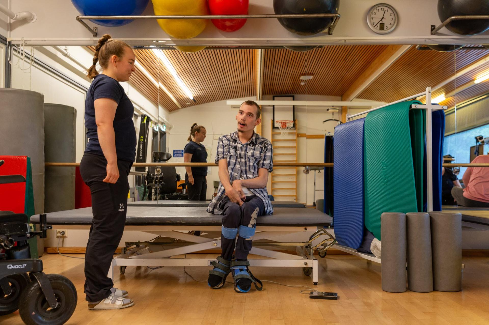En person sitter på en brits i en träningssal med vadstöd och skor med remmar. En terapeut står framför personen. Träningsredskap syns i bakgrunden.
