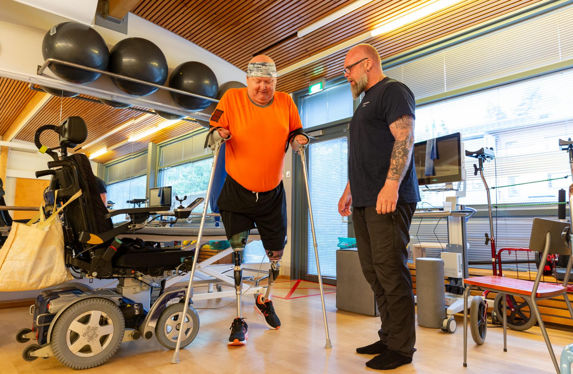 En person med två benproteser tränar gång med underarmskryckor i en rehabiliteringssal. En instruktör står bredvid. En elrullstol syns i förgrunden.