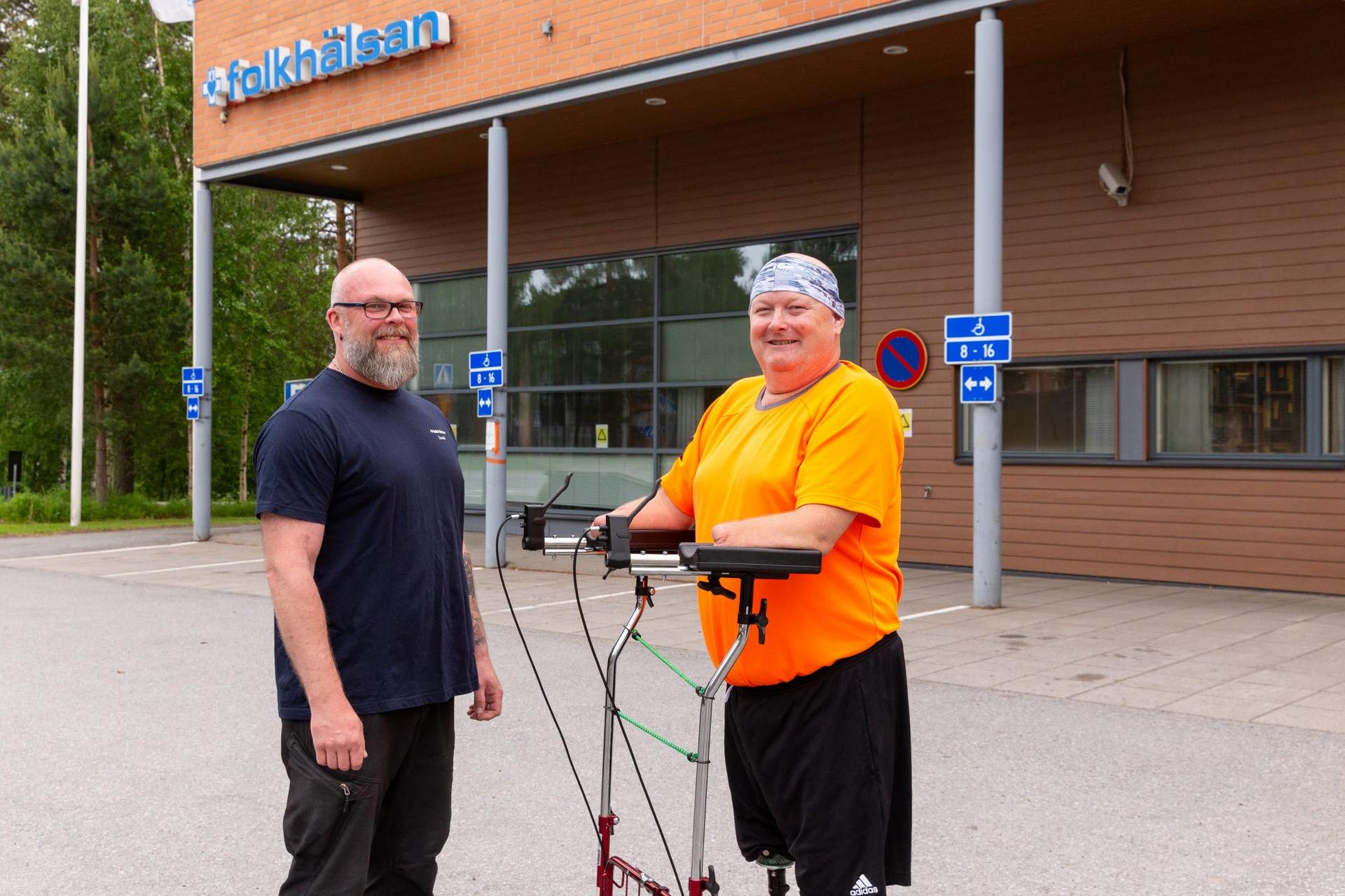Två personer står utomhus framför Folkhälsans hus i Korsholm. En person använder rollator med underarmsstöd.