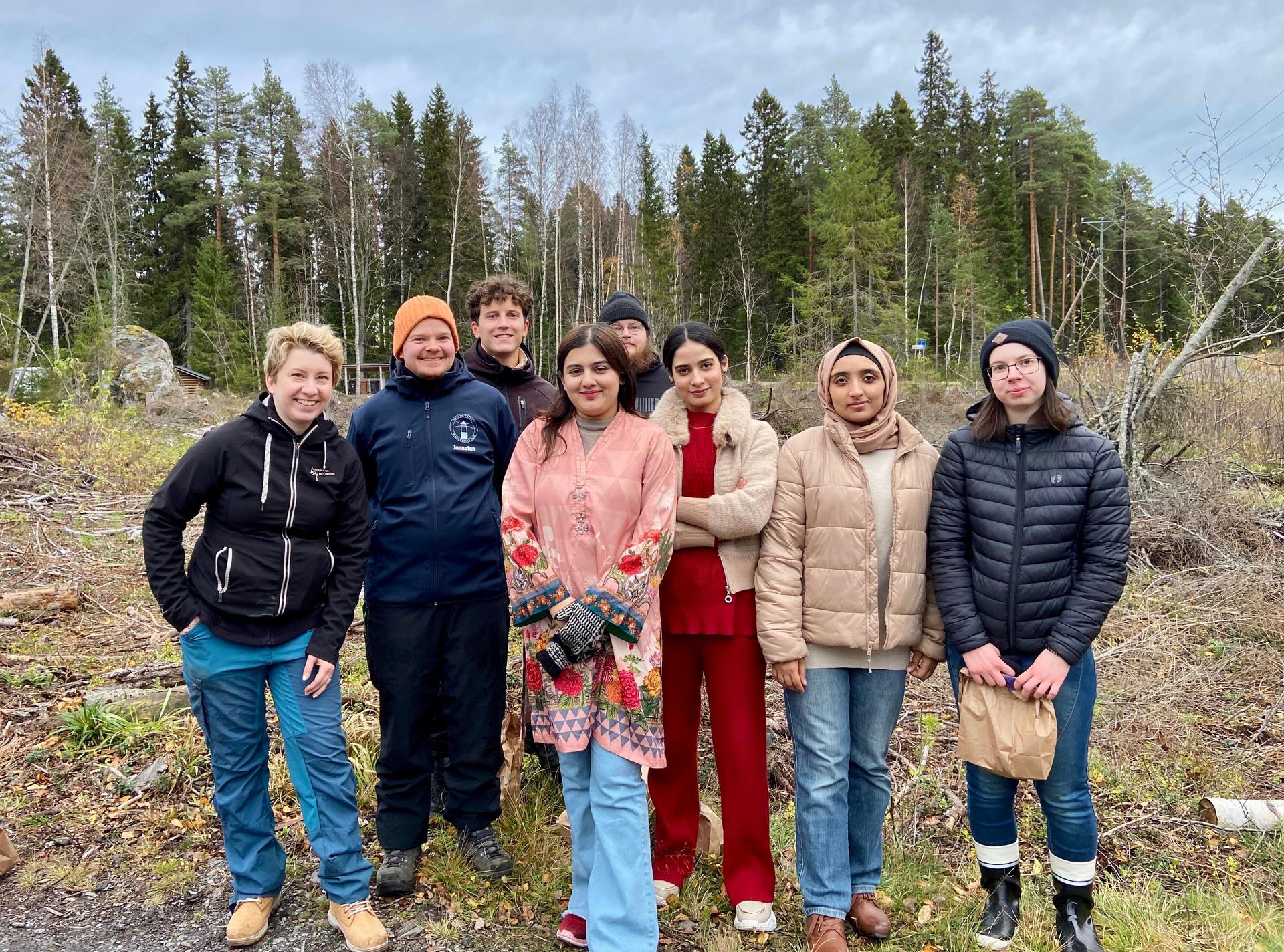 En grupp unga vuxna och en handledare står tillsammans i ett skogslandskap under en mulen dag.