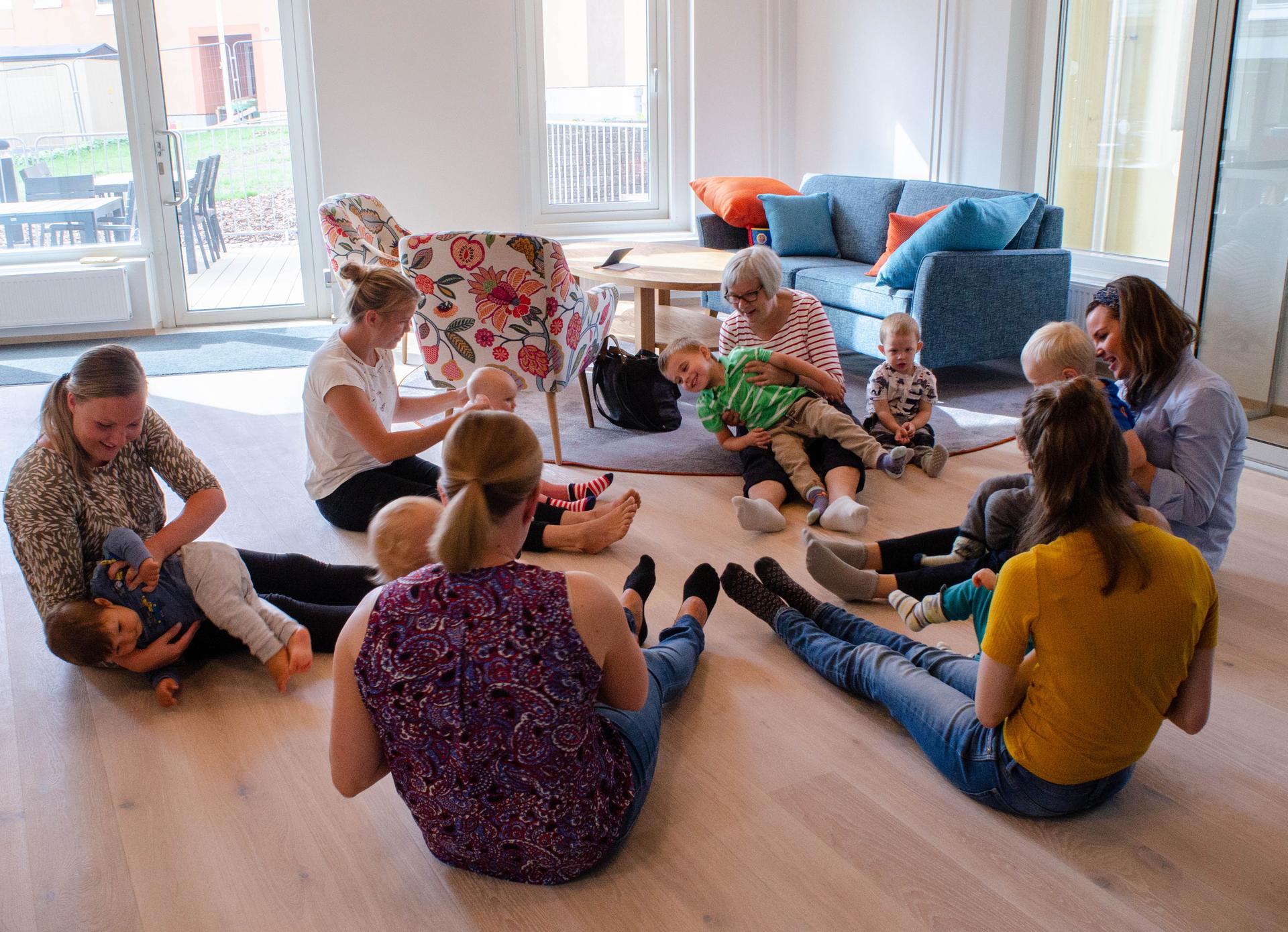 Vuxna och barn sitter i ring på golvet i ett hemtrevligt allrum med stora fönster. Barnen sitter i de vuxnas knän eller bredvid, och alla deltar i en lek eller sångstund. Miljön är ljus, med färgglada fåtöljer och soffor i bakgrunden.