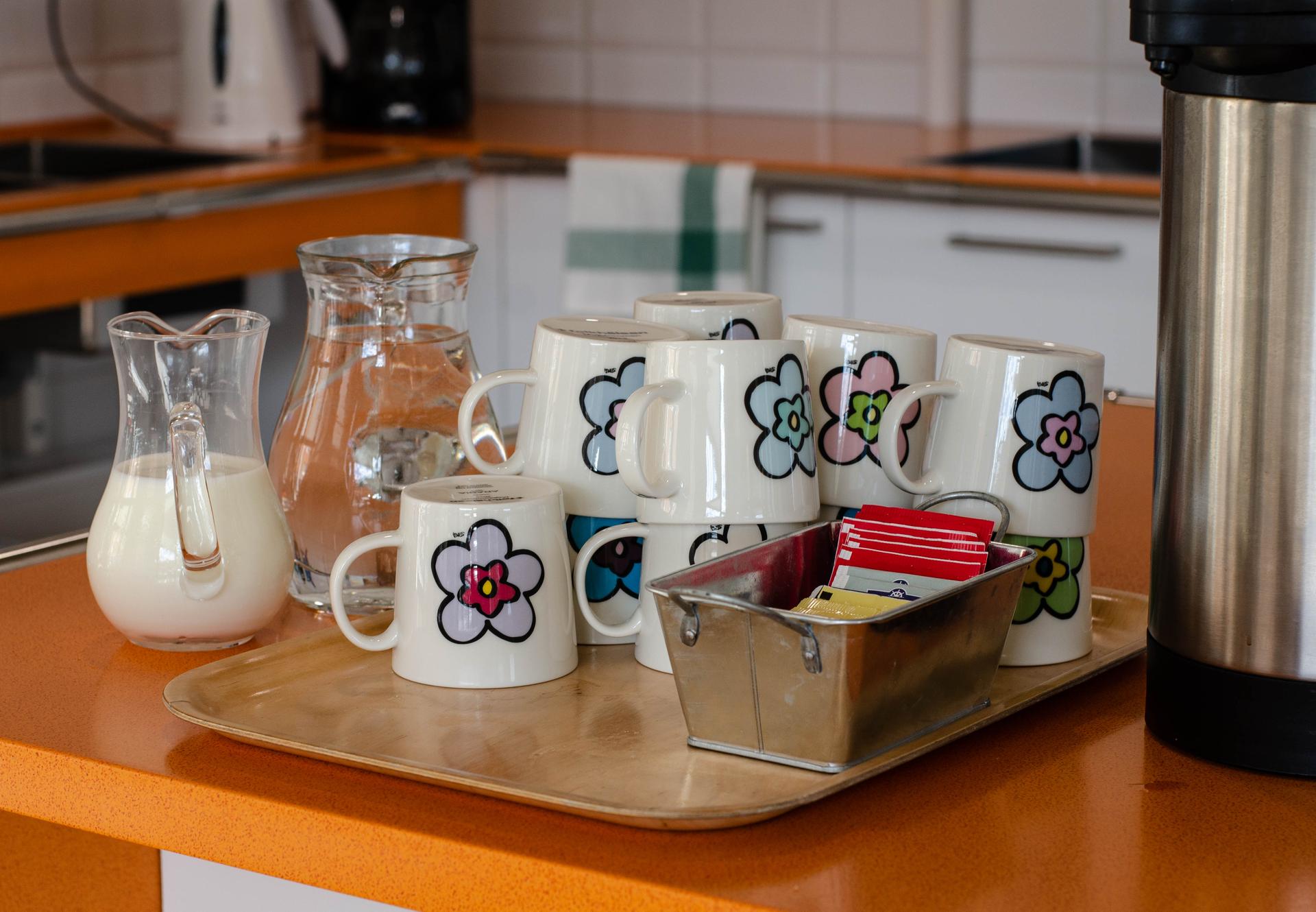Ett kök med uppdukad kaffebricka: kanna med mjölk, glaskanna med vatten, temuggar med blomdekor och ett urval av tepåsar. En termos står bredvid på en orange bänkskiva.