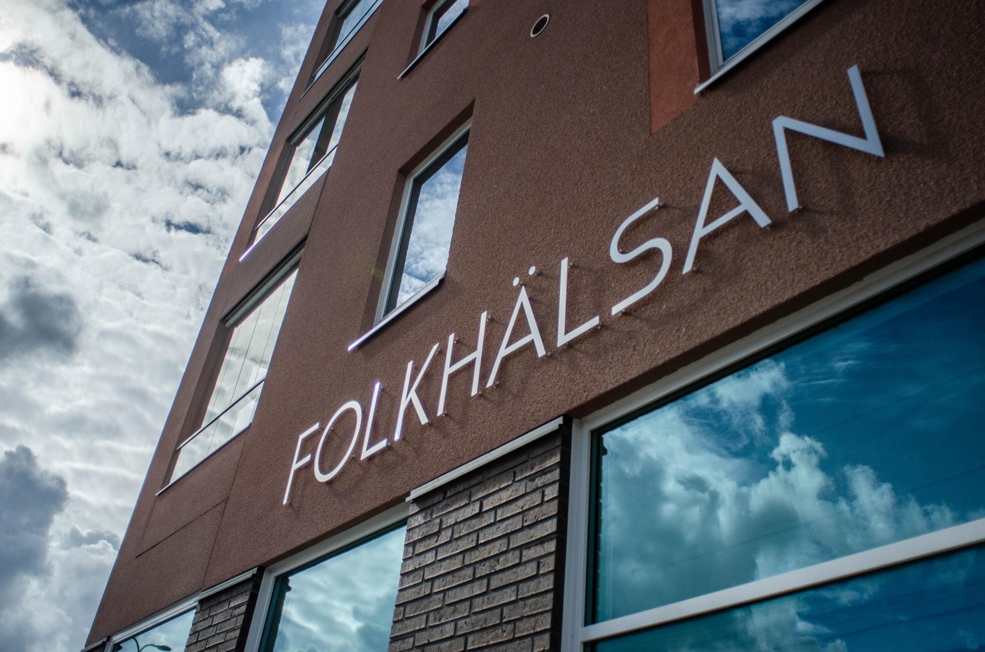 Fasadbild av ett hus med texten "FOLKHÄLSAN" i stora bokstäver. Byggnaden är brunröd och har stora fönster som speglar en dramatisk himmel med moln. Perspektivet är snett underifrån.