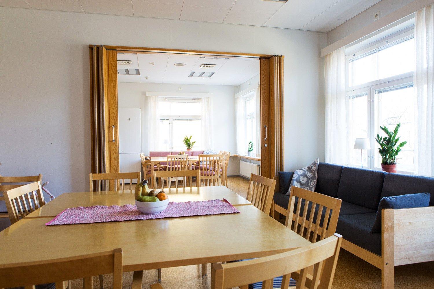 Ett ljust och hemtrevligt sällskapsrum med stora fönster, trästolar och bord. I förgrunden står ett matbord med en fruktskål. I bakgrunden syns en soffgrupp och ytterligare sittplatser.