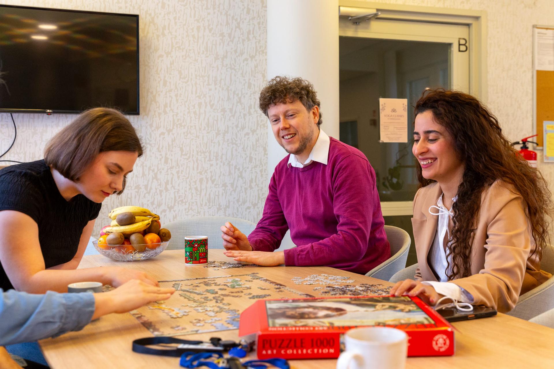 A group of people sits around a table, smiling and working together on a jigsaw puzzle. The relaxed setting includes coffee cups, a fruit bowl, and a cozy atmosphere, fostering a sense of community and collaboration.