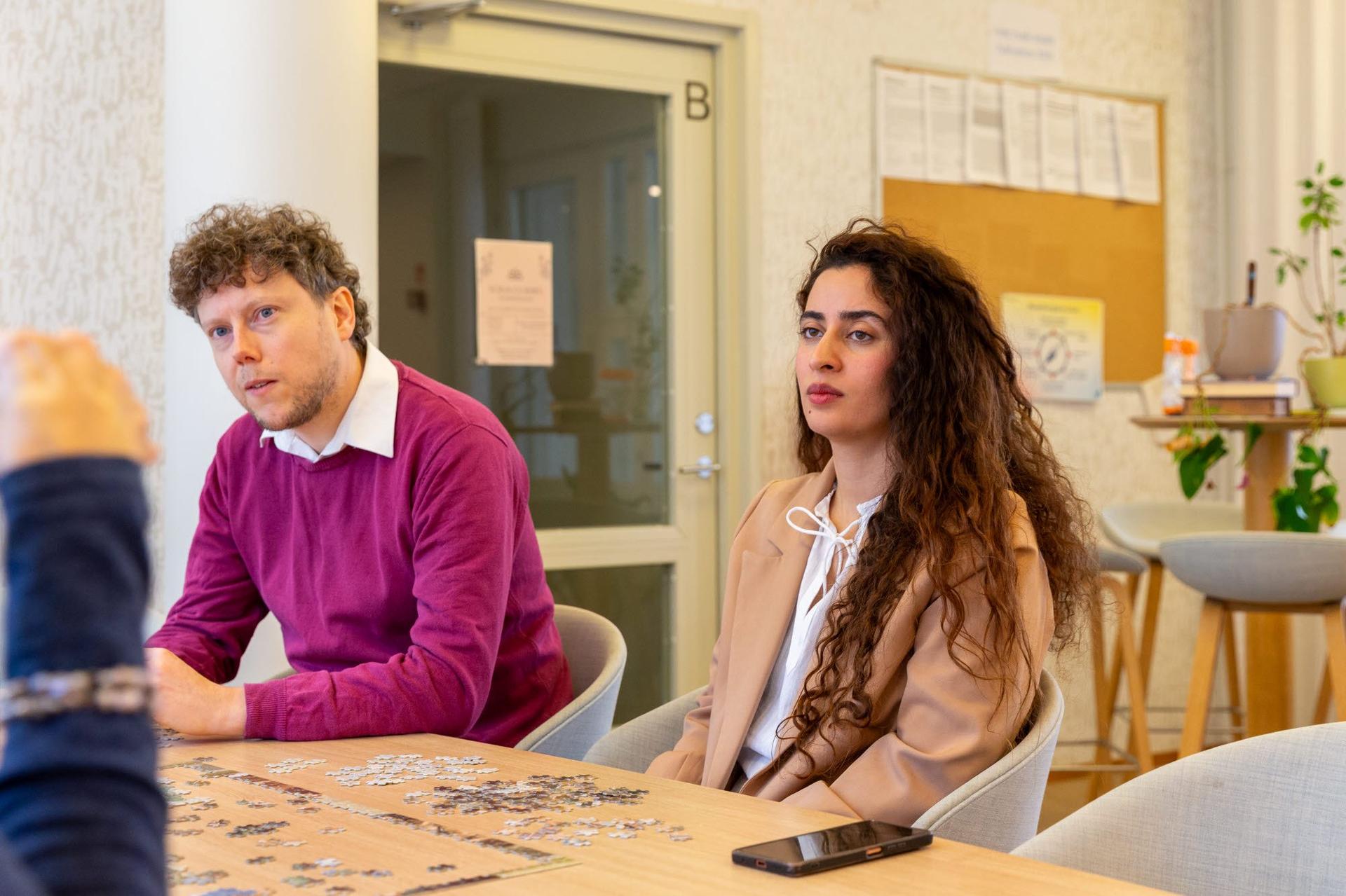 Two colleagues sit at a table in a shared office space, listening to someone out of frame while working on a jigsaw puzzle. A bulletin board and doorway are visible behind them, with a smartphone lying on the table.