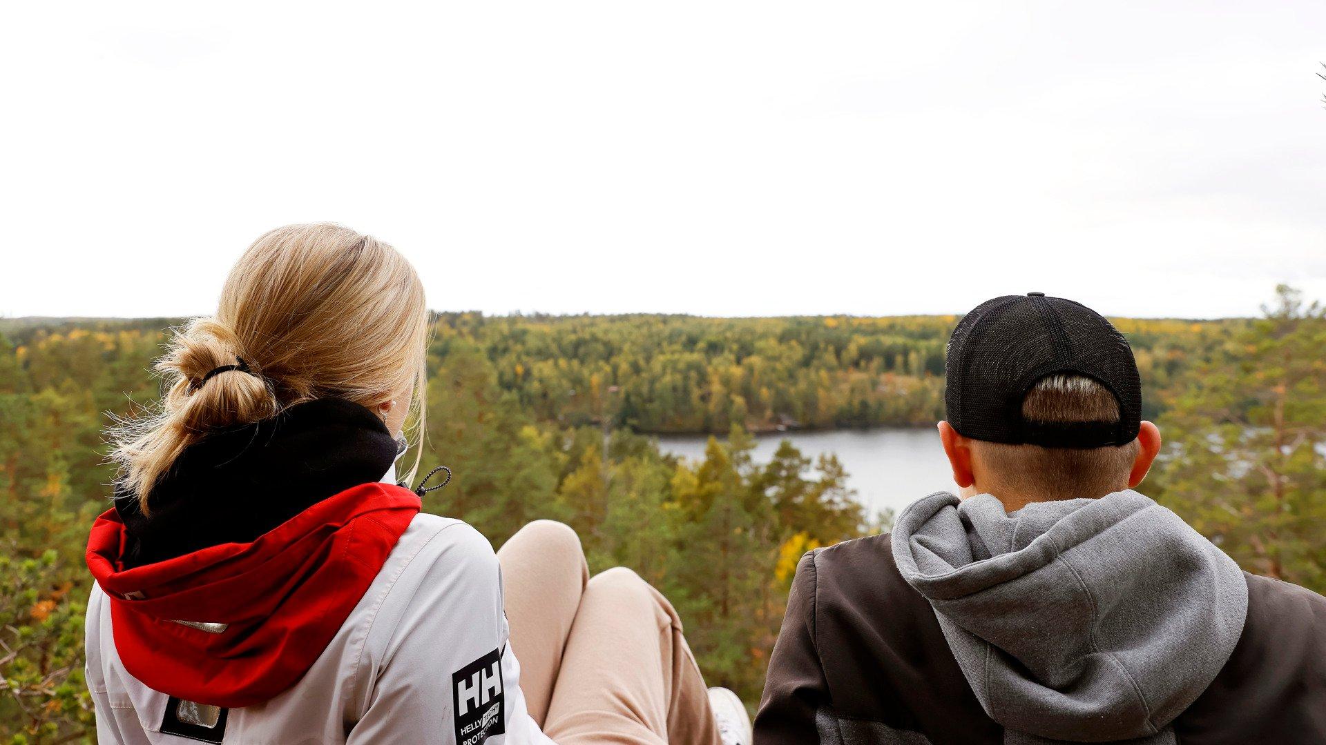 Two people sit with their backs to the camera, looking out over a forested landscape and a lake from a high viewpoint. One wears a red-and-white jacket, and the other wears a black cap and a gray hoodie.