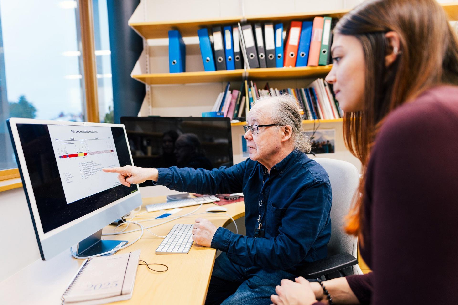 A researcher points at a computer screen displaying a data chart while discussing the results with a colleague in an office. Bookshelves and binders in the background suggest an academic or research workspace.