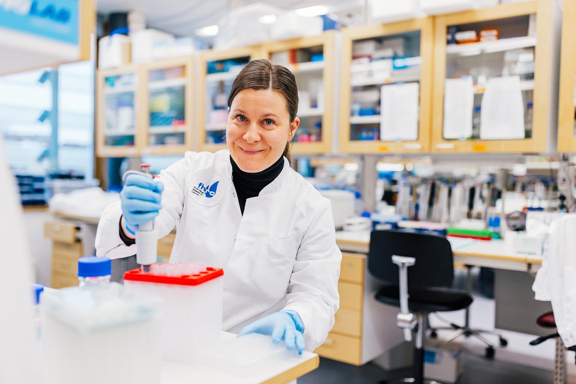 A researcher in a lab coat and blue gloves uses a pipette at a laboratory bench, smiling toward the camera. Lab cabinets and equipment are visible in the background, suggesting an active research environment.