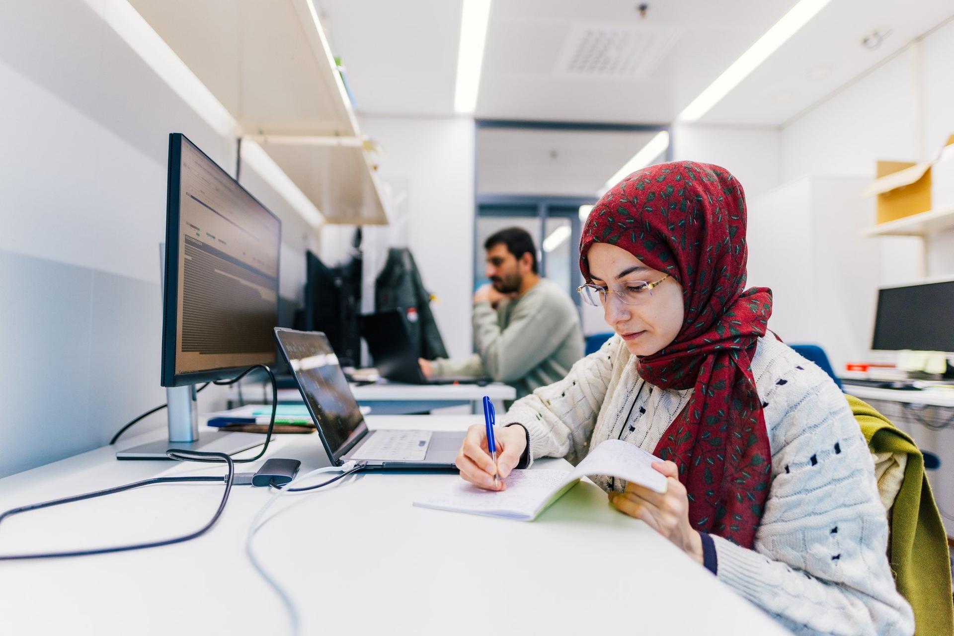A person wearing a patterned headscarf and glasses writes notes at a desk beside a laptop and a large monitor in a bright office. Another colleague works in the background, with shelves of binders and books behind them.