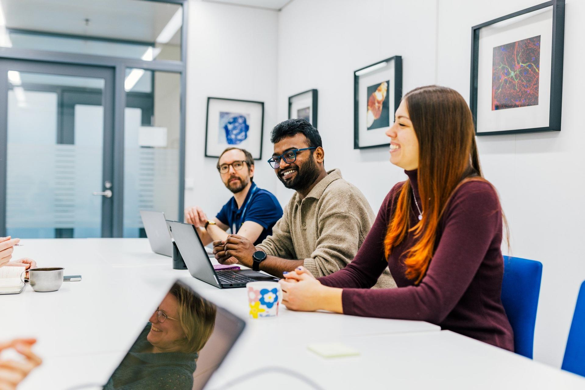 Three colleagues sit around a meeting table in a bright office, smiling and talking. Laptops and notebooks are open as they collaborate during a team discussion.