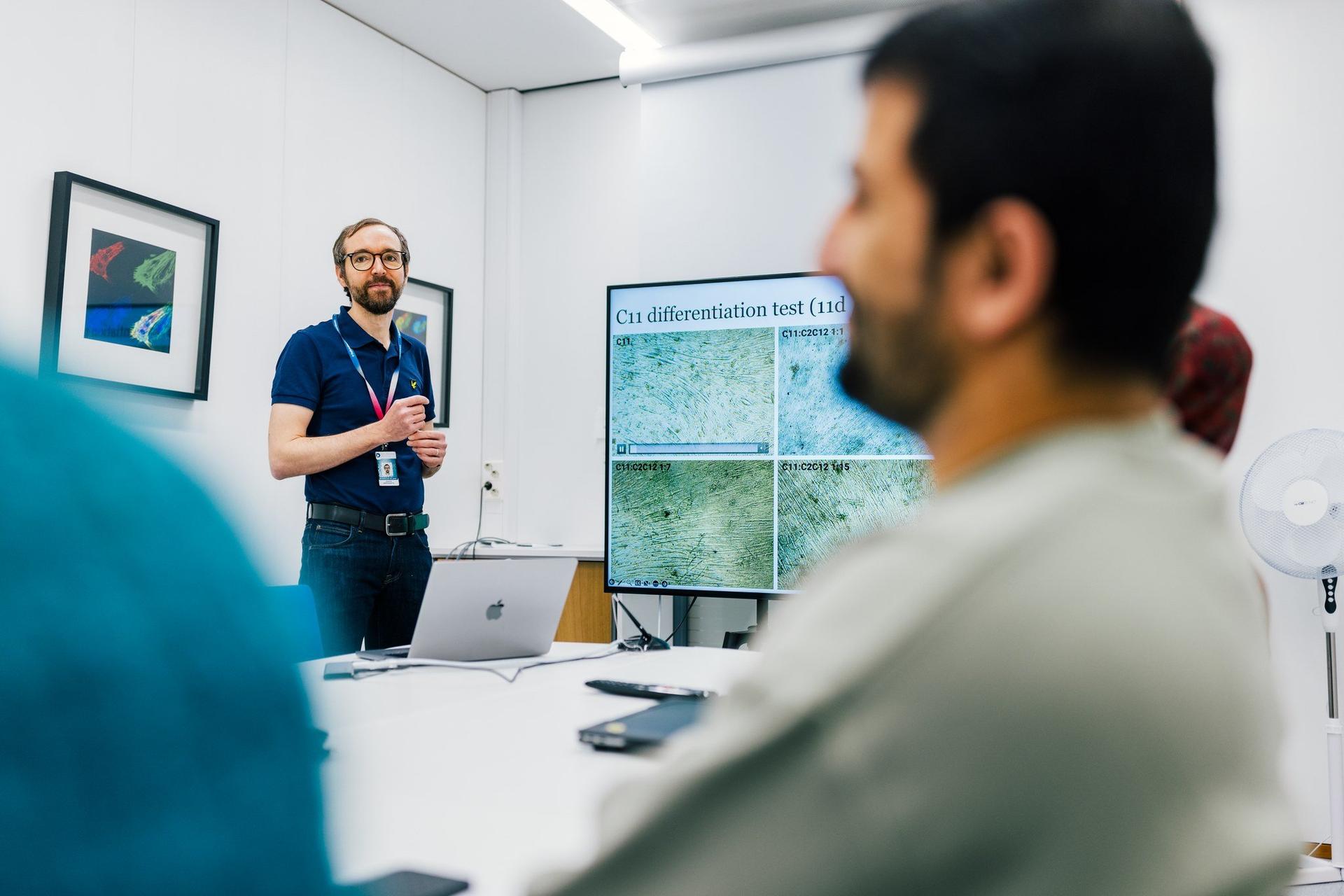 A presenter stands in a meeting room speaking to colleagues while a large screen displays microscope images labeled “C11 differentiation test (11d).” The audience is shown out of focus in the foreground, emphasizing a research or lab meeting setting.