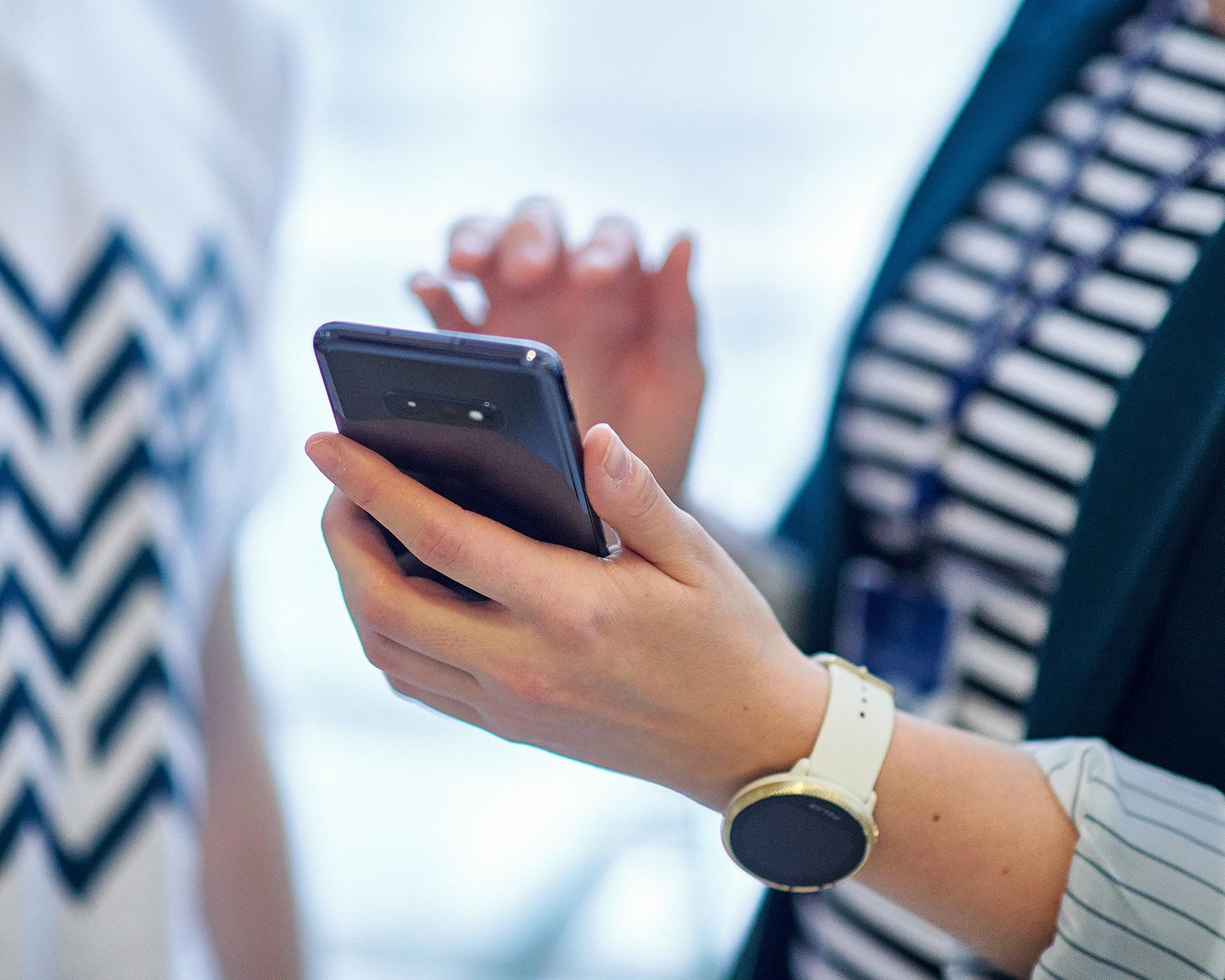 A person holds a smartphone in one hand while gesturing with the other, wearing a wristwatch and a striped shirt. The background is blurred, suggesting a conversation or workplace setting.