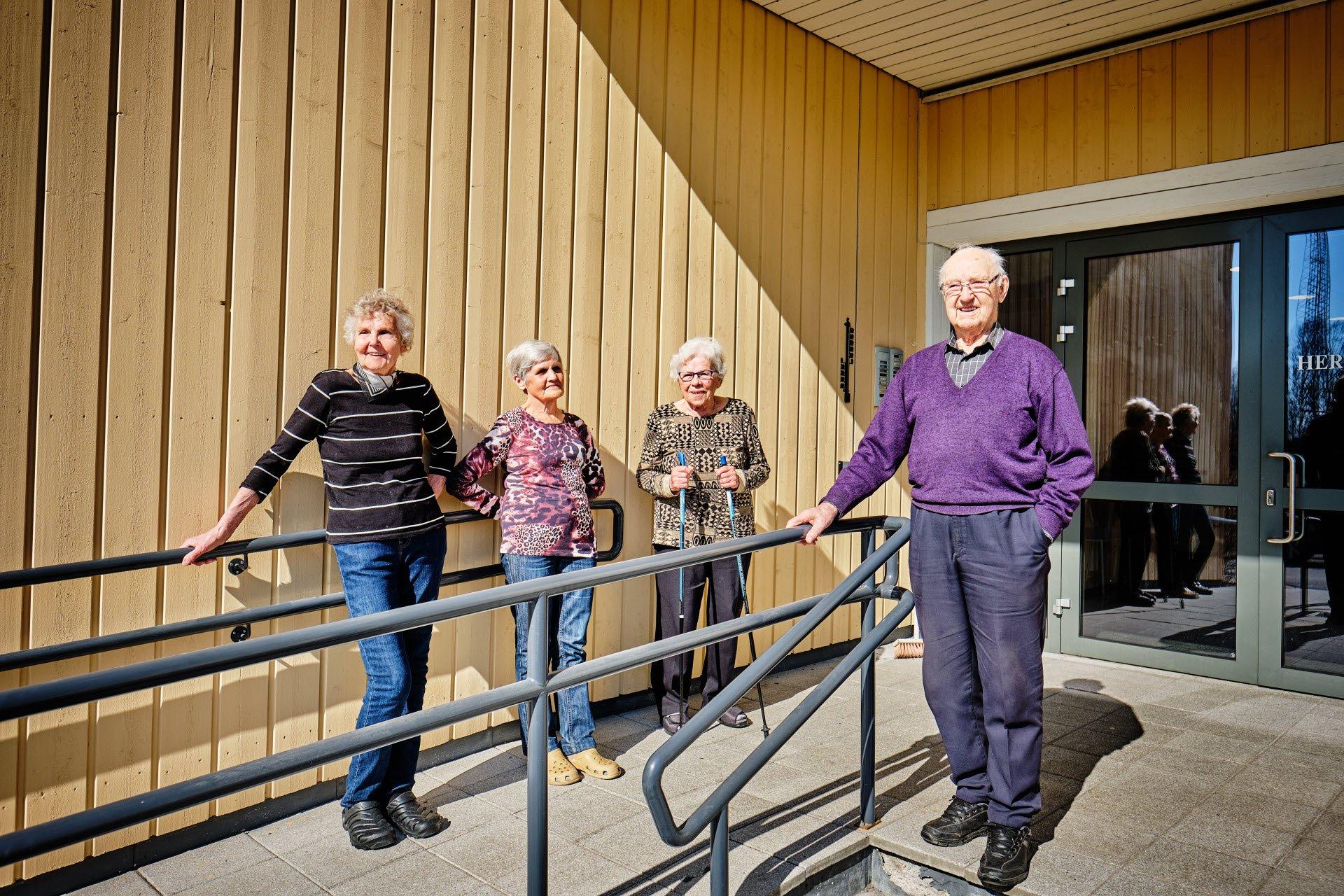 Four older adults stand outdoors along a ramp with metal railings beside a wooden building. One person stands near the entrance door while the others are positioned along the railing.