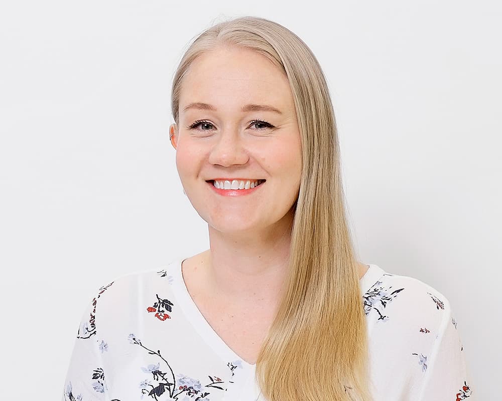A smiling woman with long blonde hair is pictured in a studio-style portrait against a white background. She is wearing a white blouse with a subtle floral pattern and is facing the camera.