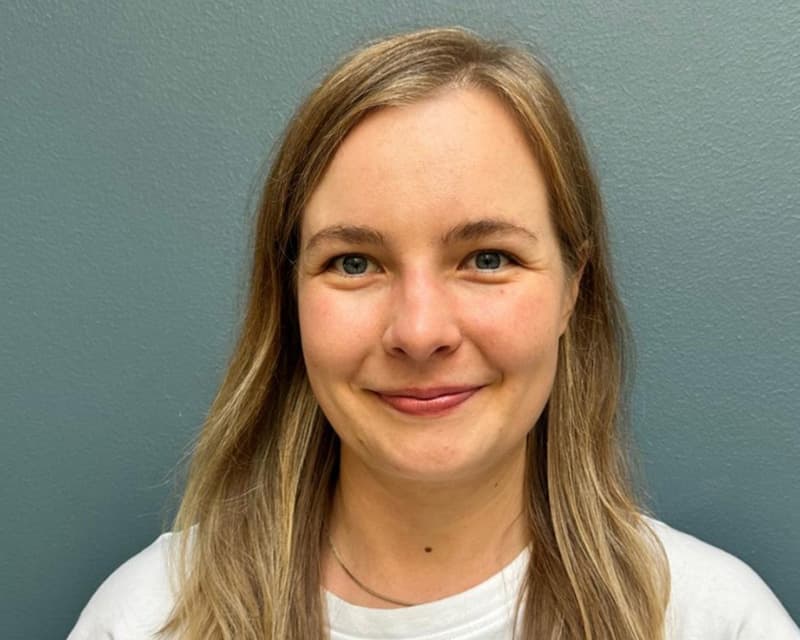 A portrait photo shows a smiling woman with long blonde hair against a muted green wall. She is facing the camera and wearing a white top.