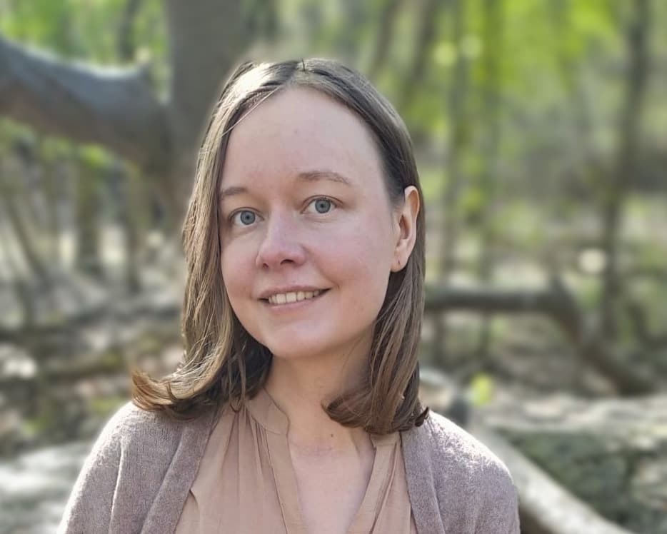 A person with shoulder-length light brown hair smiles at the camera, wearing a light top and cardigan. The background shows a softly blurred forest with green light filtering through the trees.
