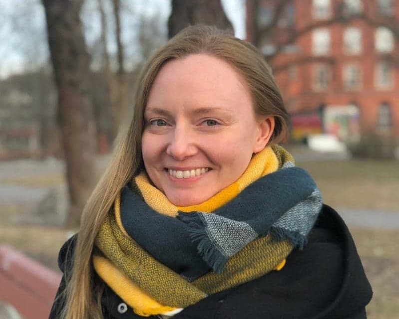 A smiling person with long blonde hair standing outdoors among trees and buildings in the background. The person is wearing a dark coat and a thick scarf in yellow, green, and gray tones.
