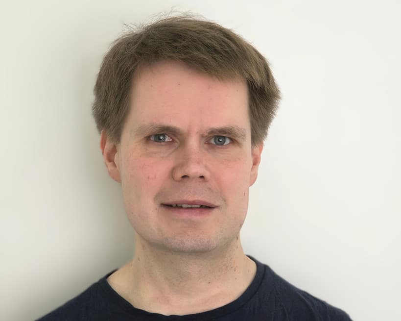 Portrait of a man with short light brown hair and blue eyes, wearing a dark shirt. He is standing against a plain light background, facing the camera with a neutral expression.