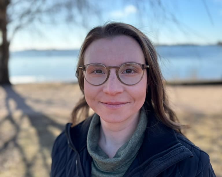A woman wearing glasses and a dark jacket smiles gently while standing outdoors on a sunny day. In the background, a calm body of water and a clear blue sky create a peaceful atmosphere.