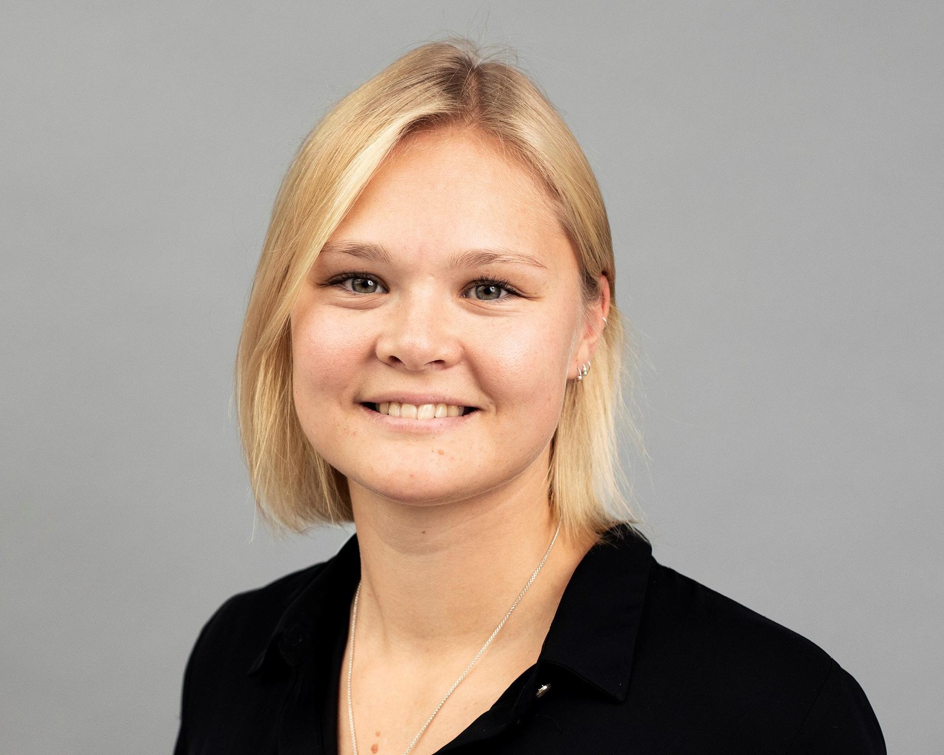 A professional headshot of a young woman with short blonde hair, wearing a black blouse and a silver necklace, smiling at the camera against a light grey background. The portrait is well-lit and framed from the chest up, creating a clean and approachable impression.