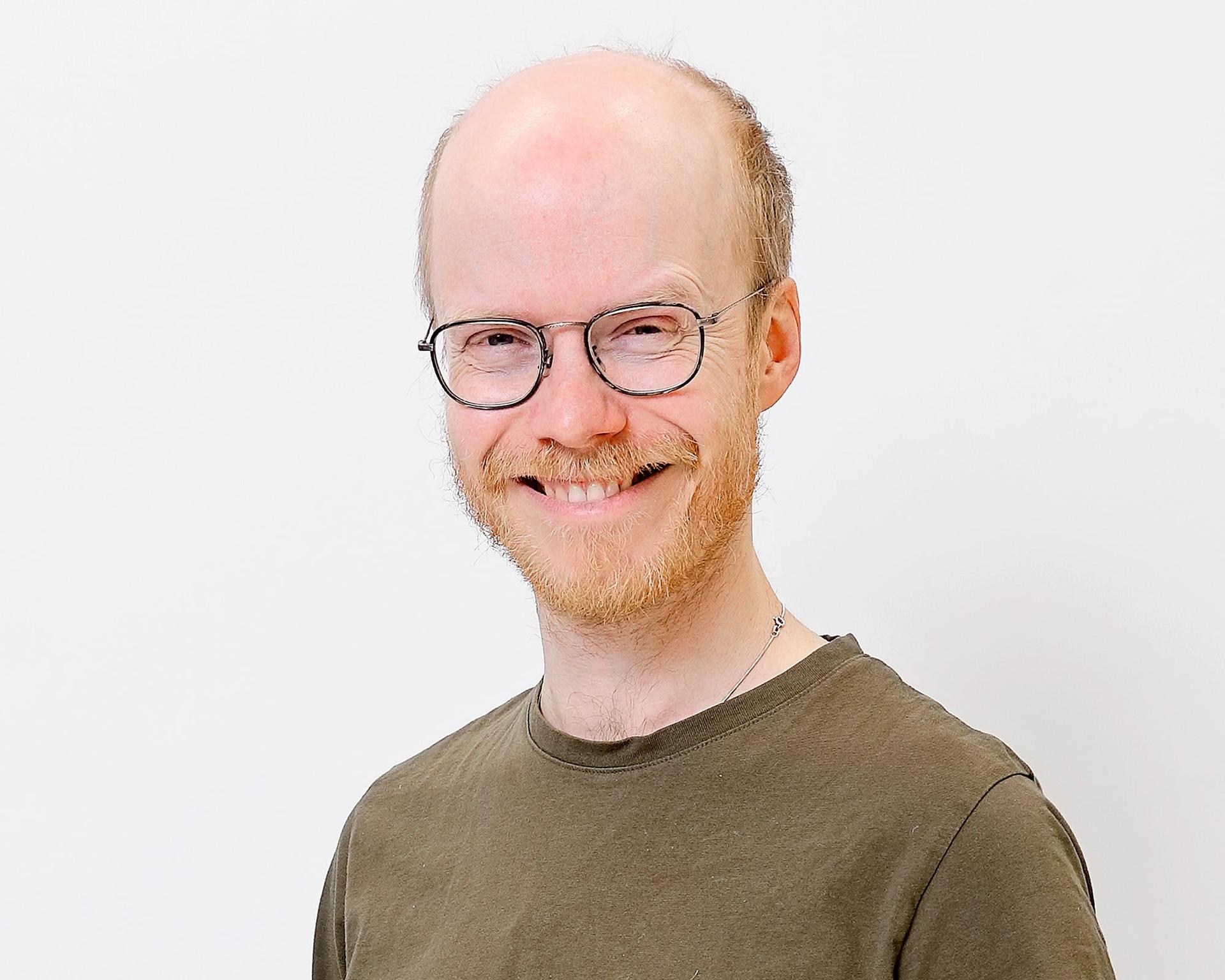 Portrait of a smiling person with glasses, a reddish beard and a green shirt against a light background. He is facing the camera with a relaxed expression.