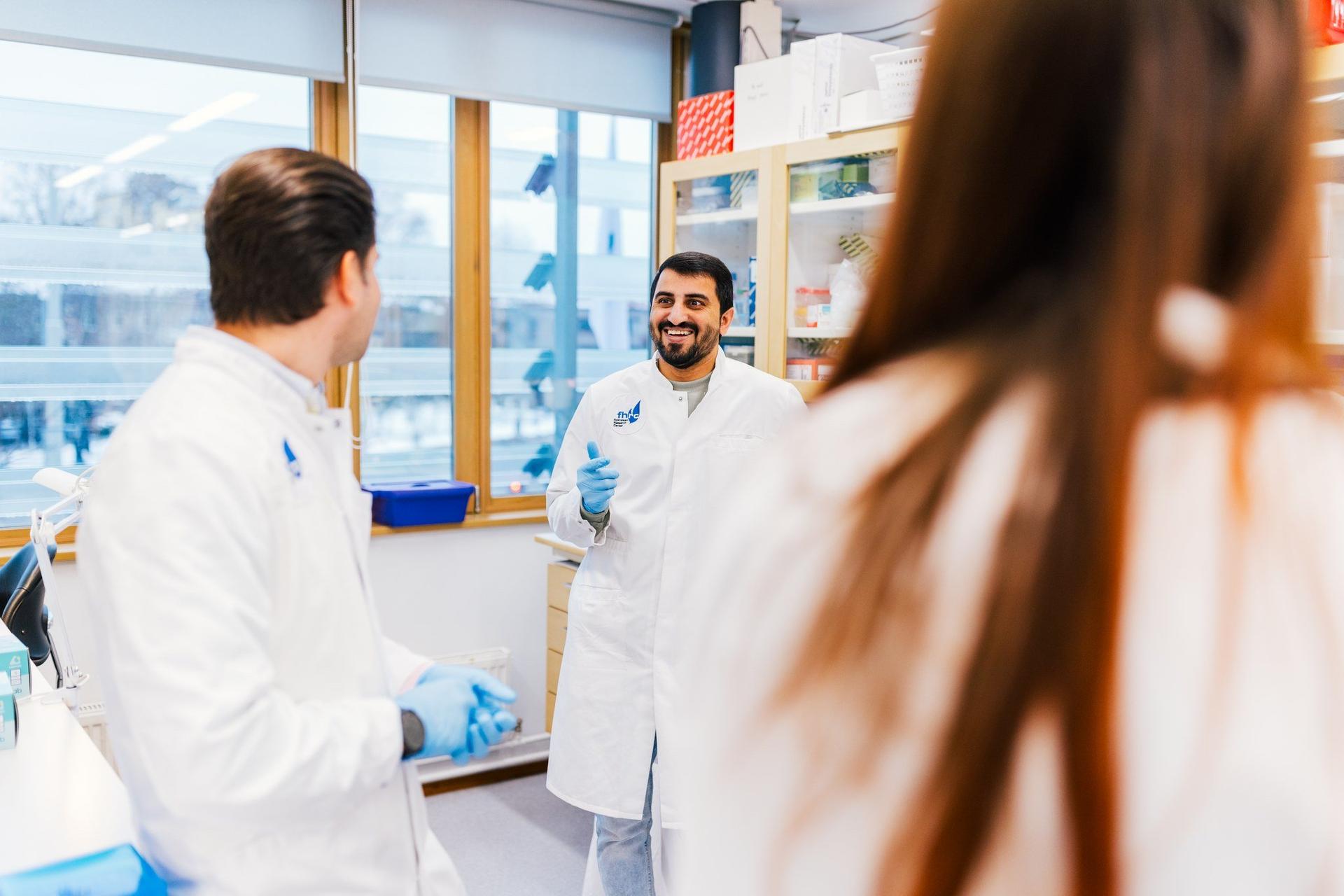 Three researchers in white lab coats and blue gloves are engaged in a conversation in a well-lit laboratory. One researcher is smiling while speaking, while the others listen attentively, surrounded by lab equipment and shelves with scientific supplies.