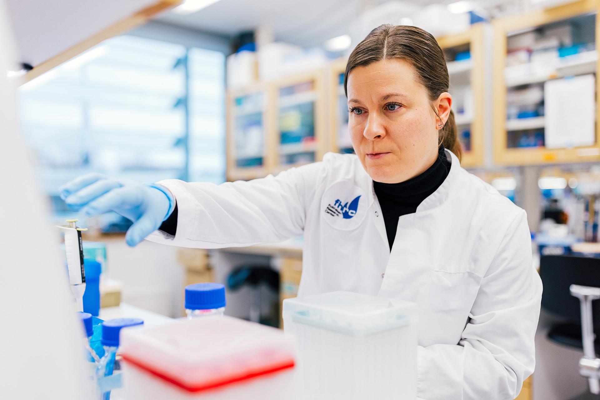 A female scientist wearing a white lab coat and blue gloves is working in a laboratory, handling scientific equipment with focus. The background features shelves filled with lab supplies and a bright window providing natural light.