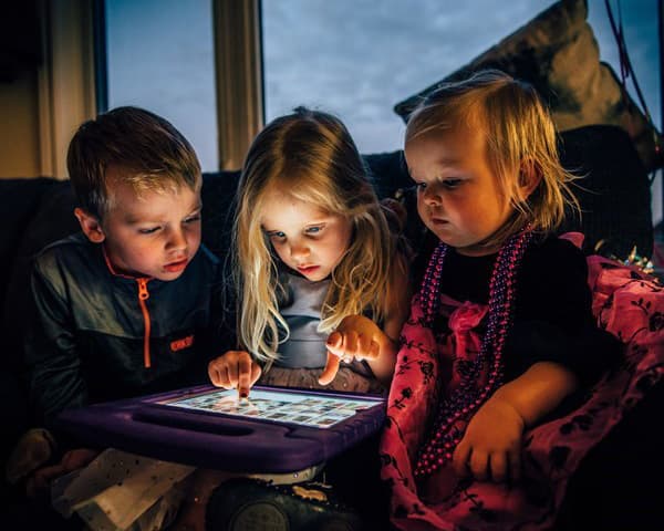 Three young children sit closely together on a couch, illuminated by the glow of a tablet screen. They are focused on the device, with one child actively touching the screen while the others watch intently.