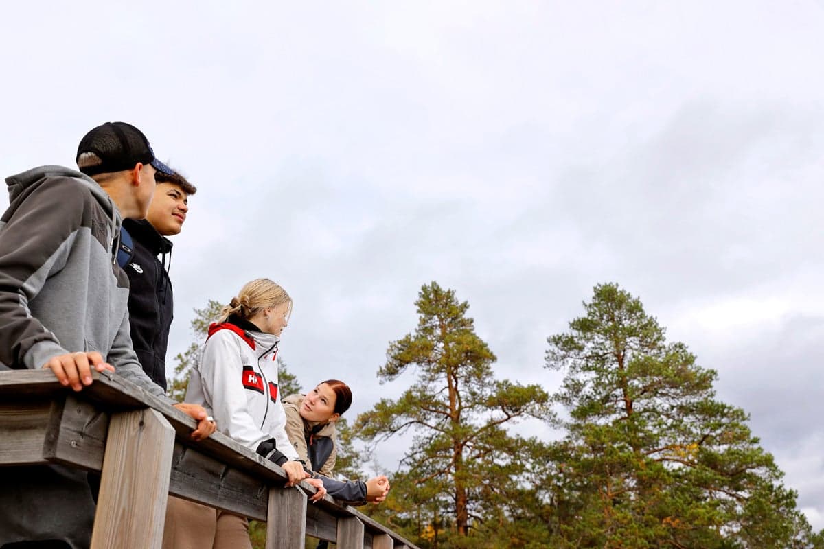 Four young people leaning against a wooden railing outdoors, with pine trees and a cloudy sky in the background. One person is wearing a white jacket with red and black details, while the others are dressed in dark outdoor clothing.