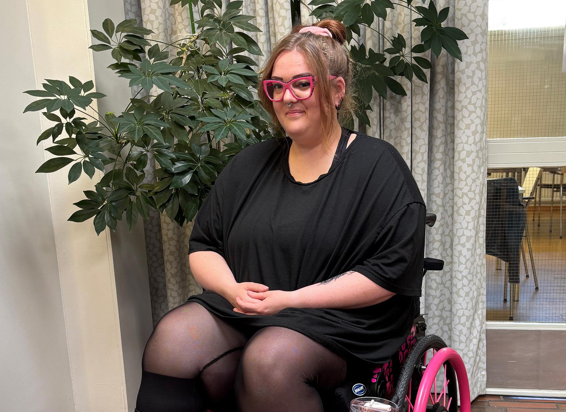 A person with pink glasses sits in a wheelchair, smiling, with hands folded in their lap. A leafy indoor plant and patterned curtains are in the background.