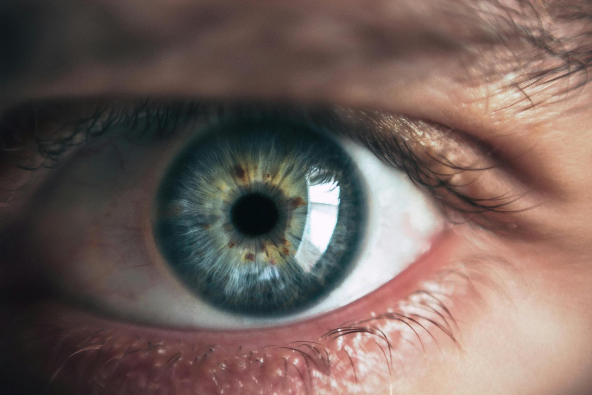 Close-up photo of a human eye with a blue-green iris and dark pupil. Eyelashes and surrounding skin are visible in soft focus.