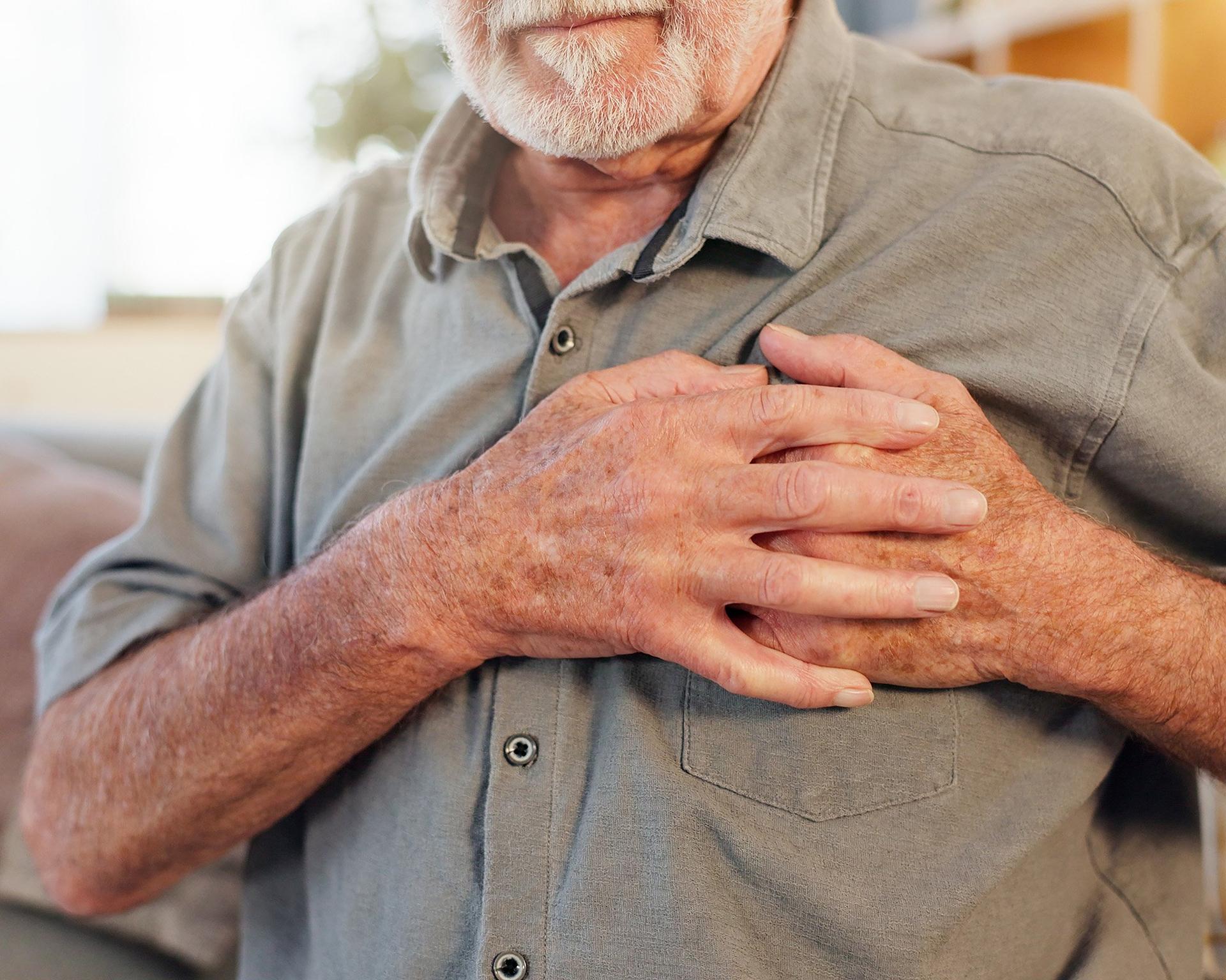 An older person in a grey shirt holds both hands against their chest. The image shows a close-up of the upper body with the face partially out of frame.