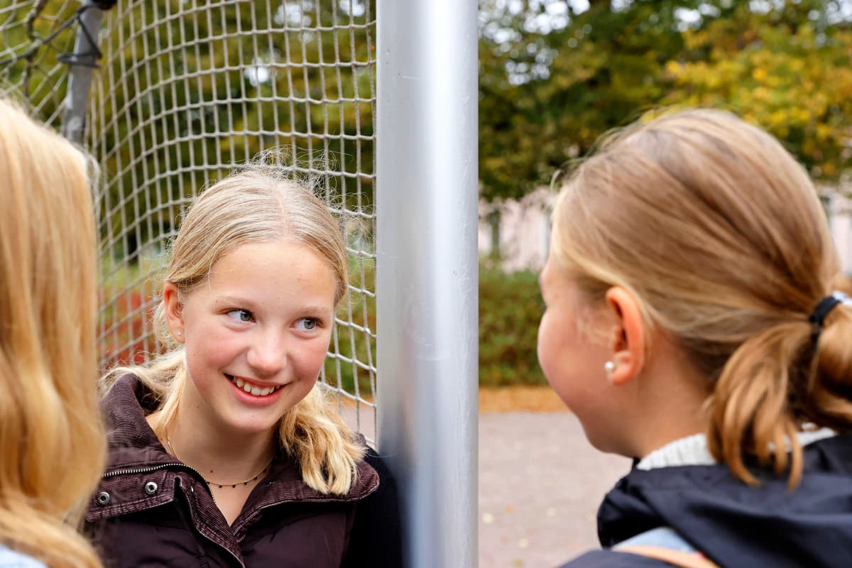 Three girls are standing by a soccer goal outdoors, talking to each other. One of the girls is smiling as she looks at her friends.