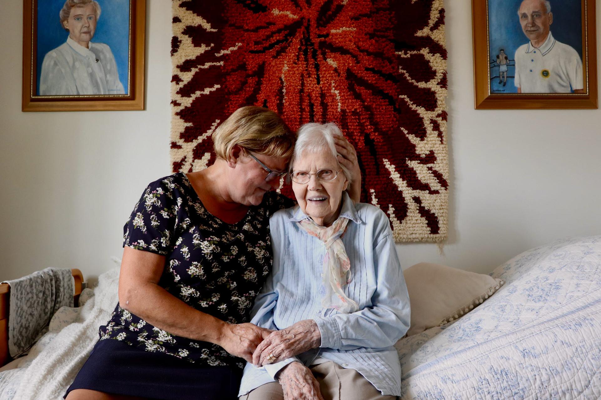 An elderly woman sits on a bed, smiling gently while holding hands with a middle-aged woman who leans in with affection. The cozy room is decorated with a woven wall hanging and framed portraits, creating a warm and personal atmosphere.