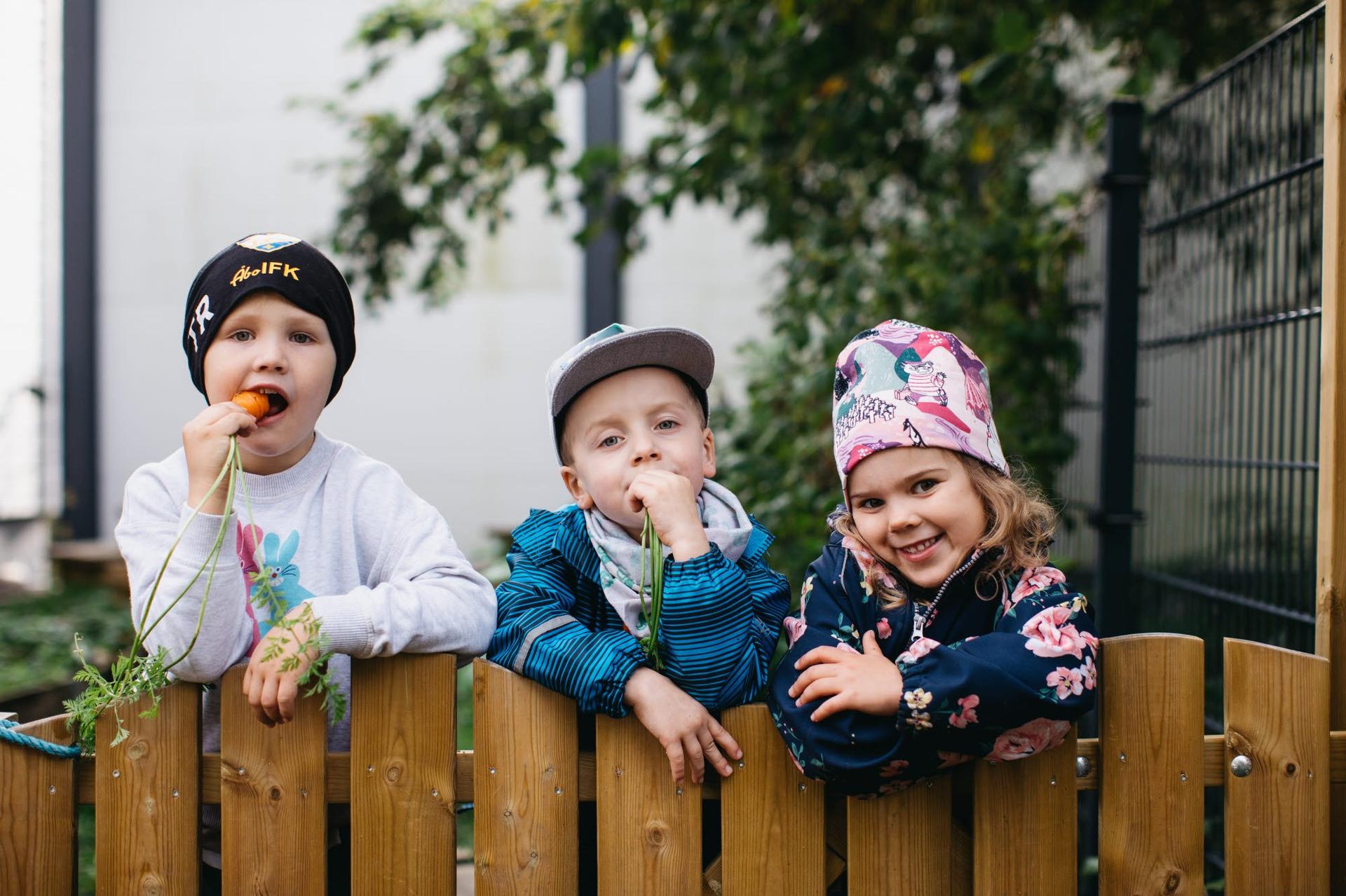 Three young children are leaning over a wooden fence, smiling and playfully eating freshly picked carrots. They are dressed in warm clothing and hats, with a garden and greenery in the background.
