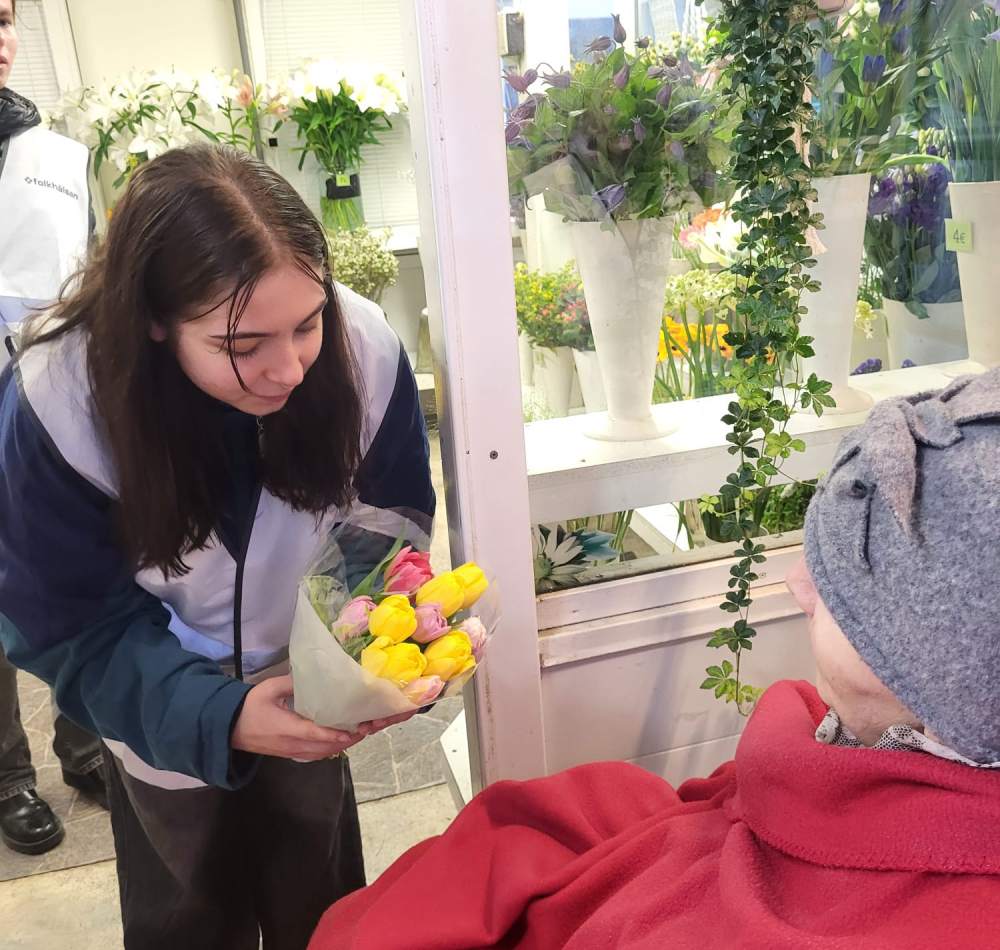 En &auml;ldre person i rullstol f&aring;r en bukett blommor av en f&ouml;rs&auml;ljerska i en blombutik.