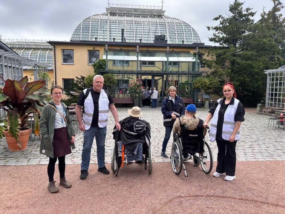Två äldre i rullstol, tre vårdare och en anhörig står framför ingången till Botaniska trädgården.
