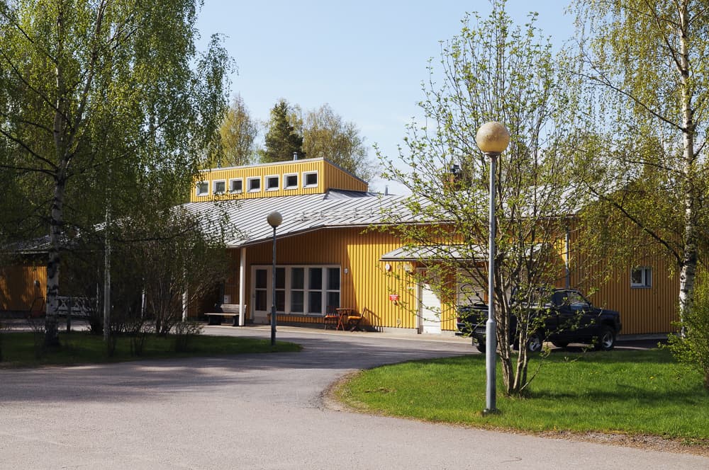 Folkhälsans servicehem i Korsholm, en gul träbyggnad med silverfärgat plåttak, omgiven av grönskande björkar och en välkomnande infart. En svart bil är parkerad vid byggnaden, och uteplatsen har utemöbler för de boende och besökare.