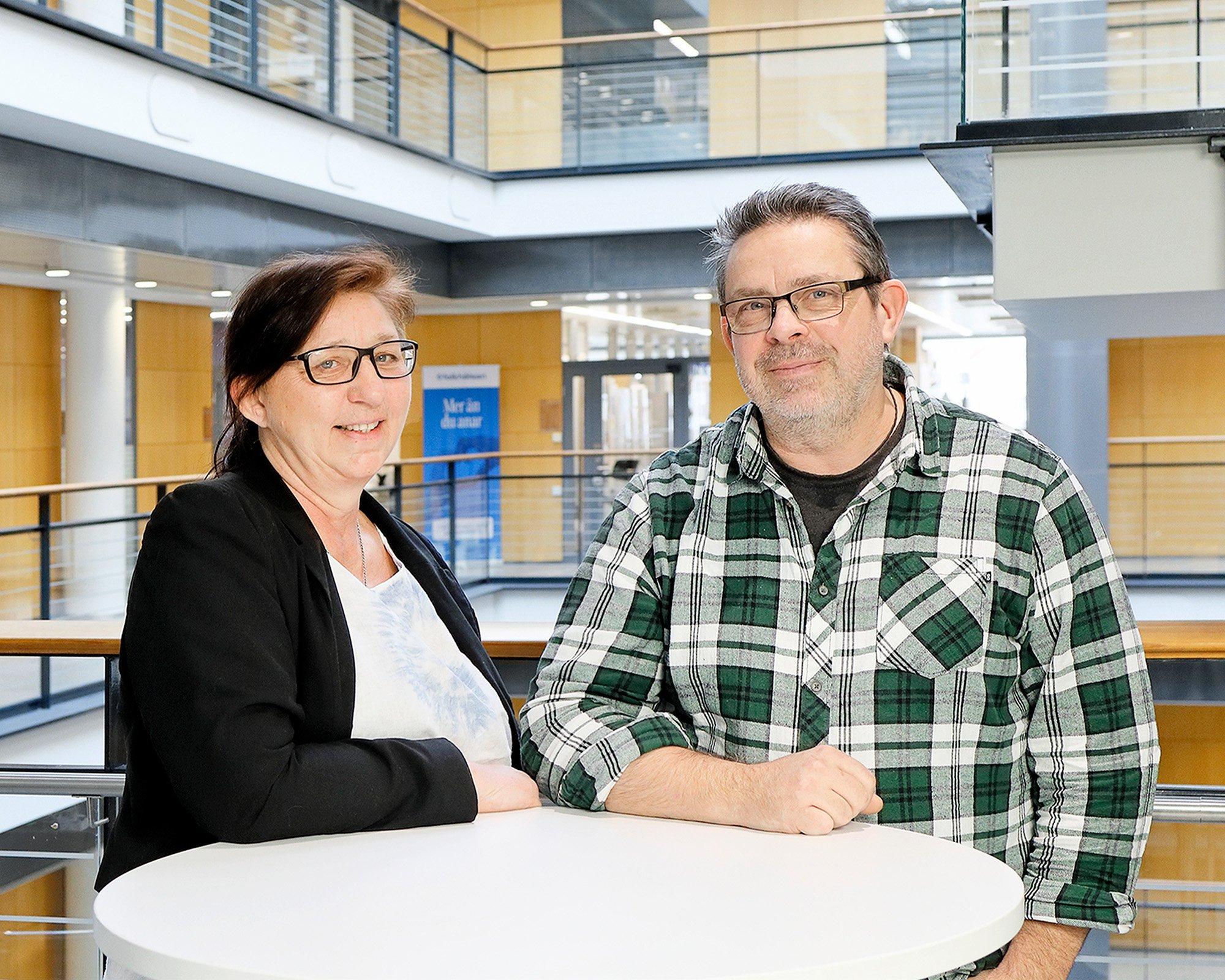 Two persons are standing indoors by a round white table in a modern, brightly lit building with glass railings and wood-paneled walls. Both are smiling at the camera – one wears a dark blazer and glasses, the other a green checkered shirt and glasses.