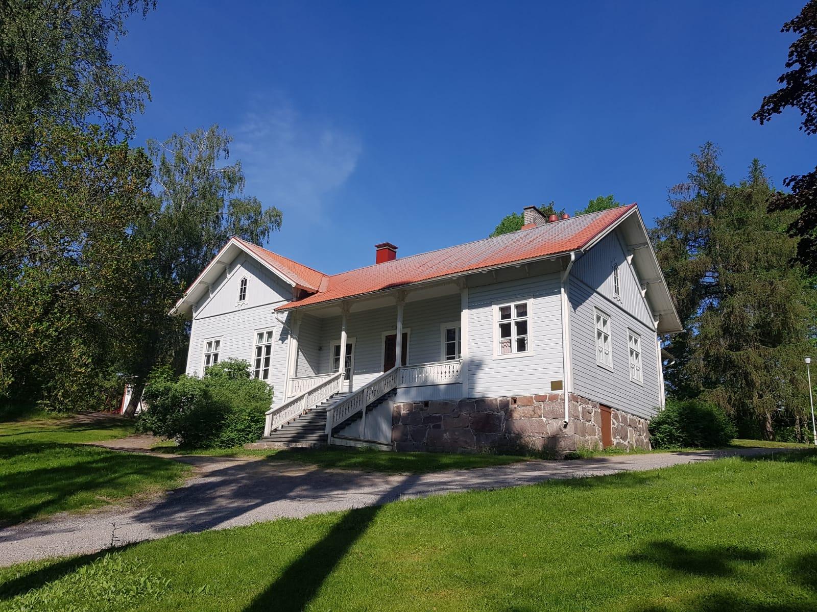En traditionell trävilla med ljusgrå fasad och rött plåttak, omgiven av grönskande träd och en välskött gräsmatta. Huset har en stentrappa som leder upp till en liten veranda. En klarblå himmel och soligt väder förstärker den idylliska atmosfären.