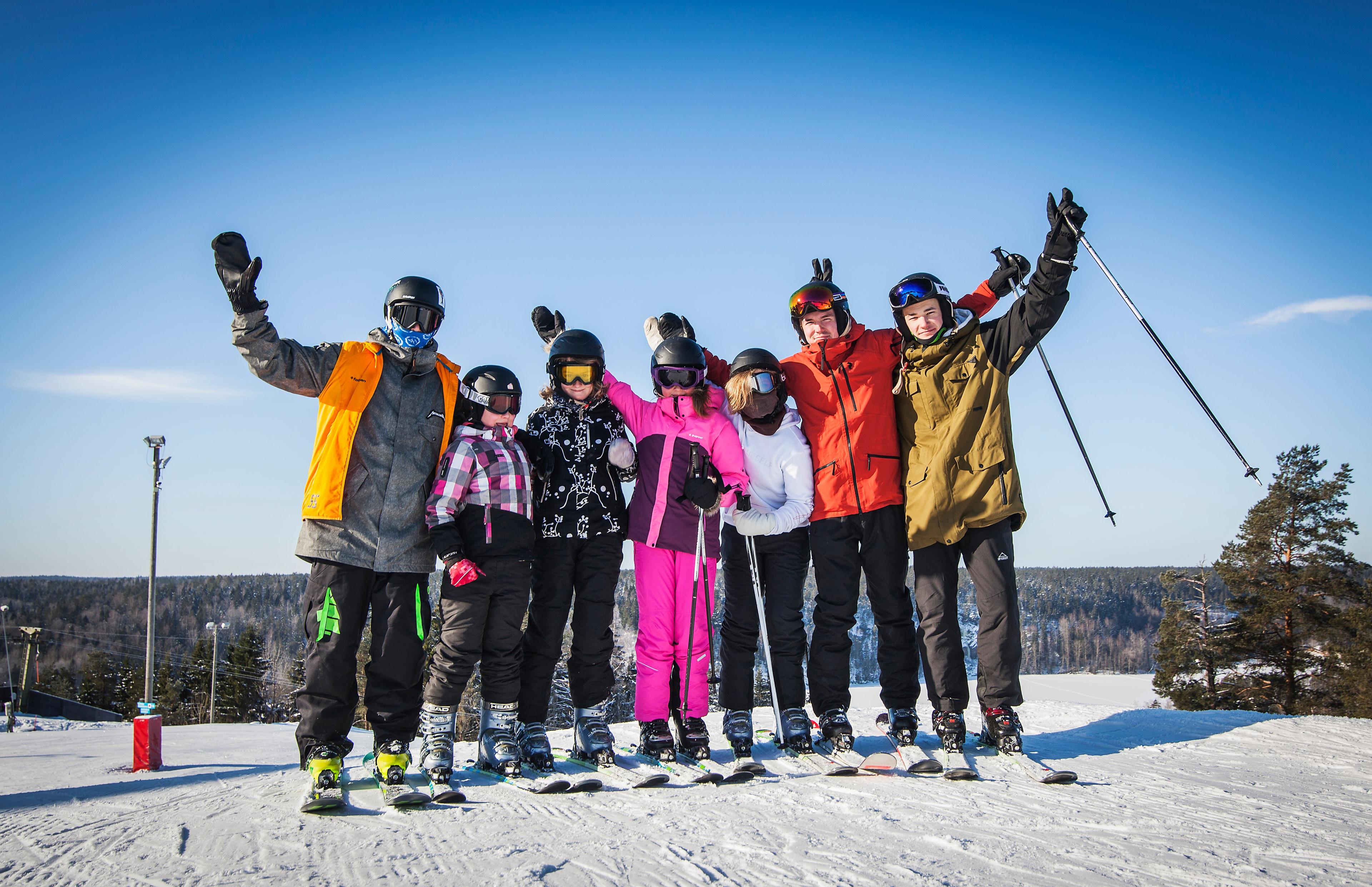 En grupp barn och unga står på skidor i en rad i en snöig skidbacke. De bär hjälmar och skidkläder i olika färger och håller armarna upp i luften. I bakgrunden syns träd och himmel.
