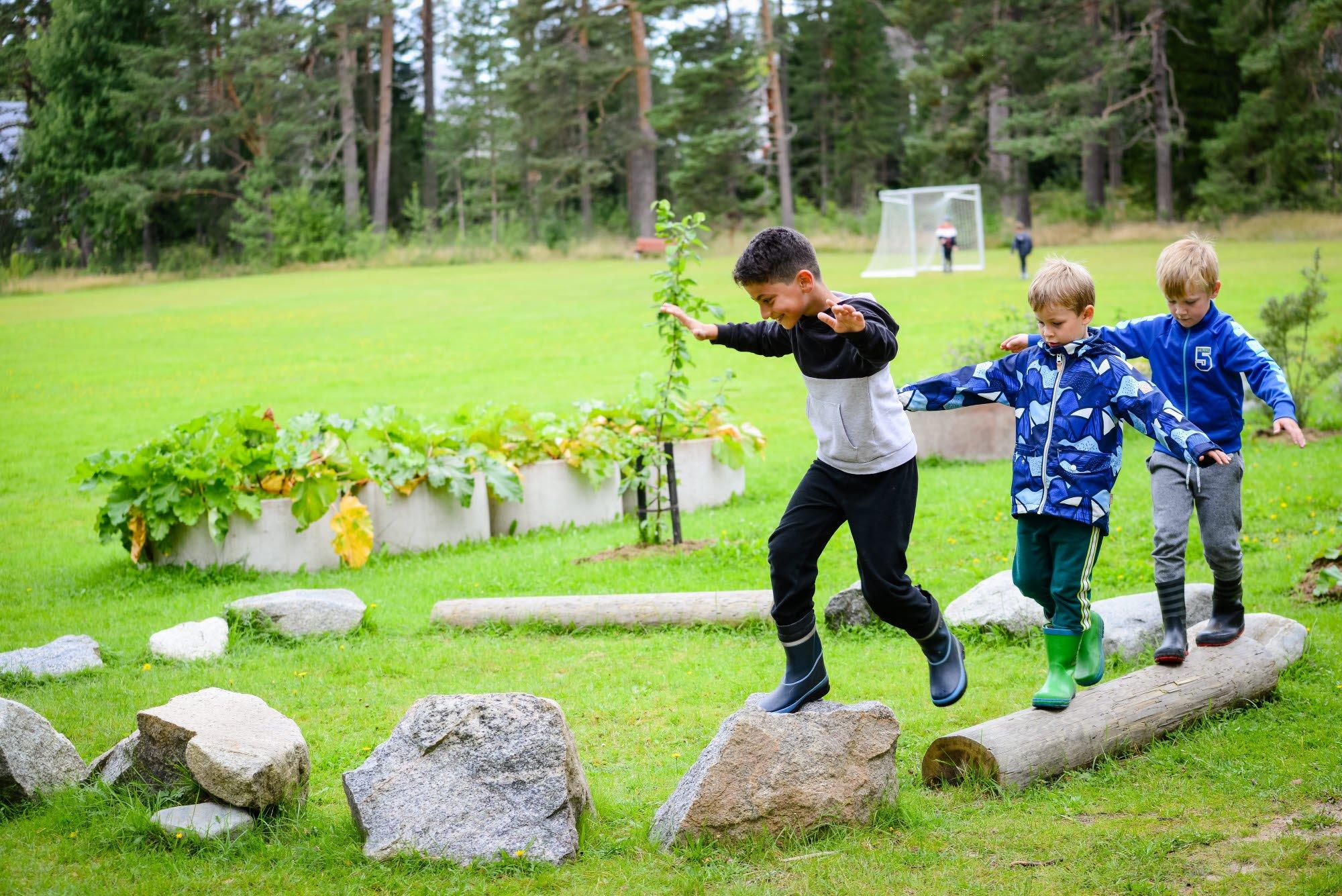 Tre barn balanserar på stenar och stockar i en grönskande utomhusmiljö, medan de leker och utmanar sin balans.