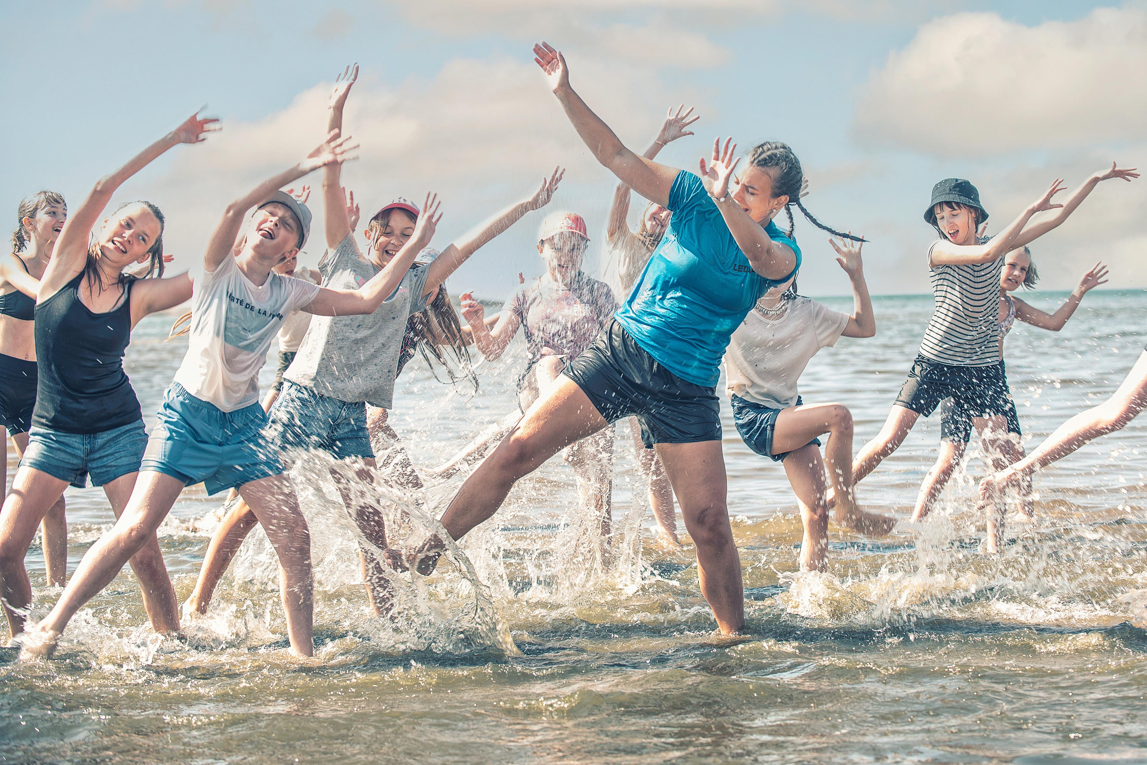Barn och en vuxen person står i grunt vatten och rör sig med armarna uppsträckta. Vatten stänker runt dem.