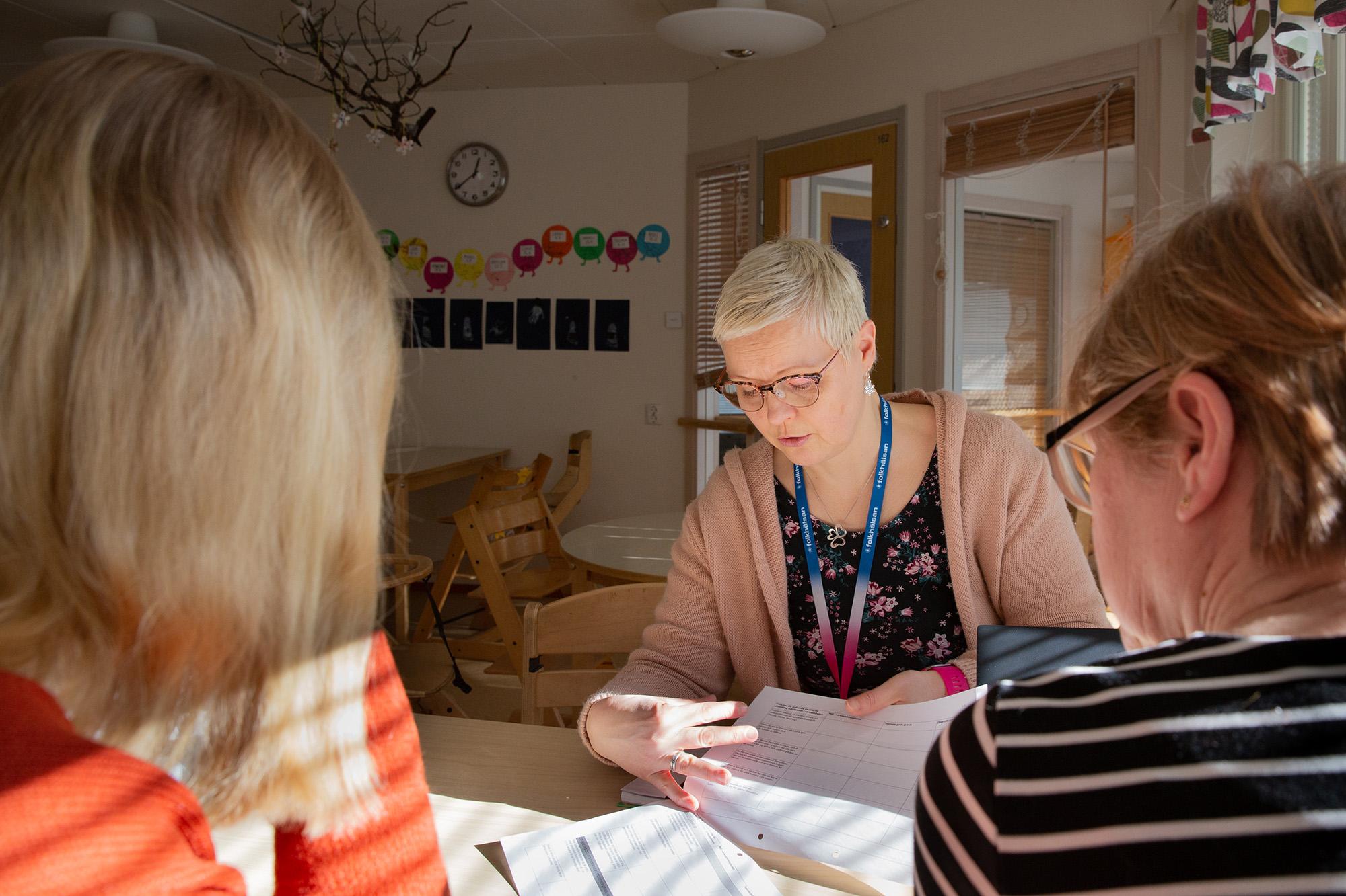 Speciallärare Nina Knutars sitter vid ett bord i en pedagogisk miljö och går igenom dokument tillsammans med två kollegor. Solen lyser in genom fönstret och skapar en varm atmosfär. Bakgrunden visar en förskolemiljö med barnteckningar och pedagogiska dekorationer på väggen.