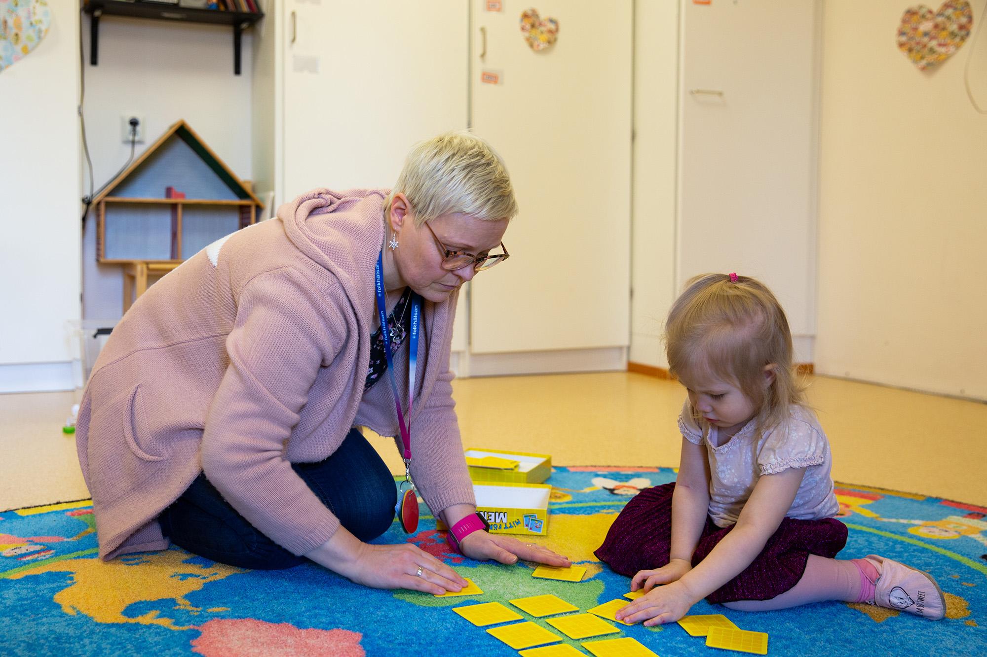 Speciallärare Nina Knutars sitter på en färgglad matta och spelar memory med ett barn i en pedagogisk miljö. Barnet är fokuserat på korten, och Nina engagerar sig i leken. Bakgrunden visar en lekhörna med ett dockhus och väggdekorationer.