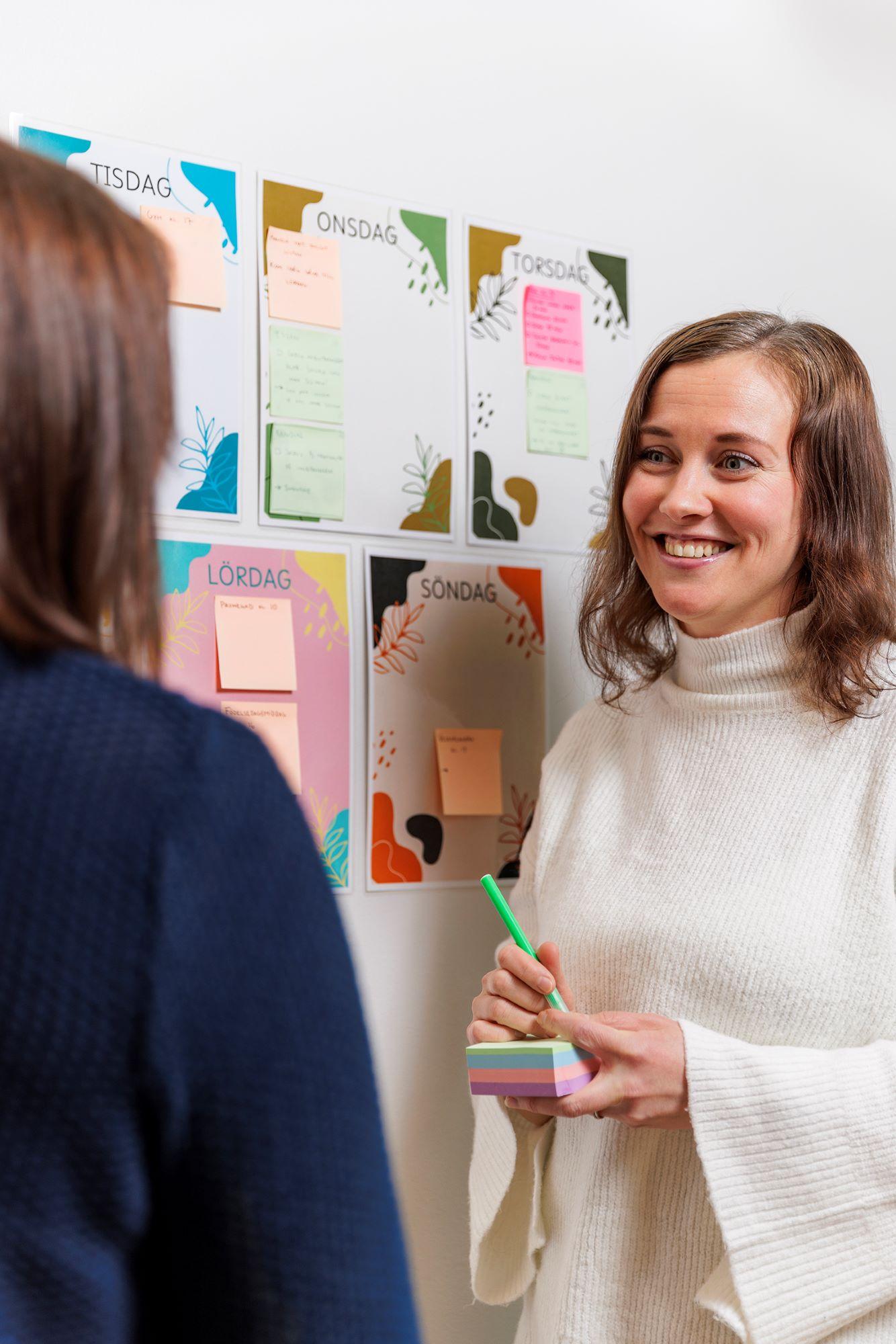 Två kvinnor står framför en vägg med post it-lappar och skriver på lapparna.