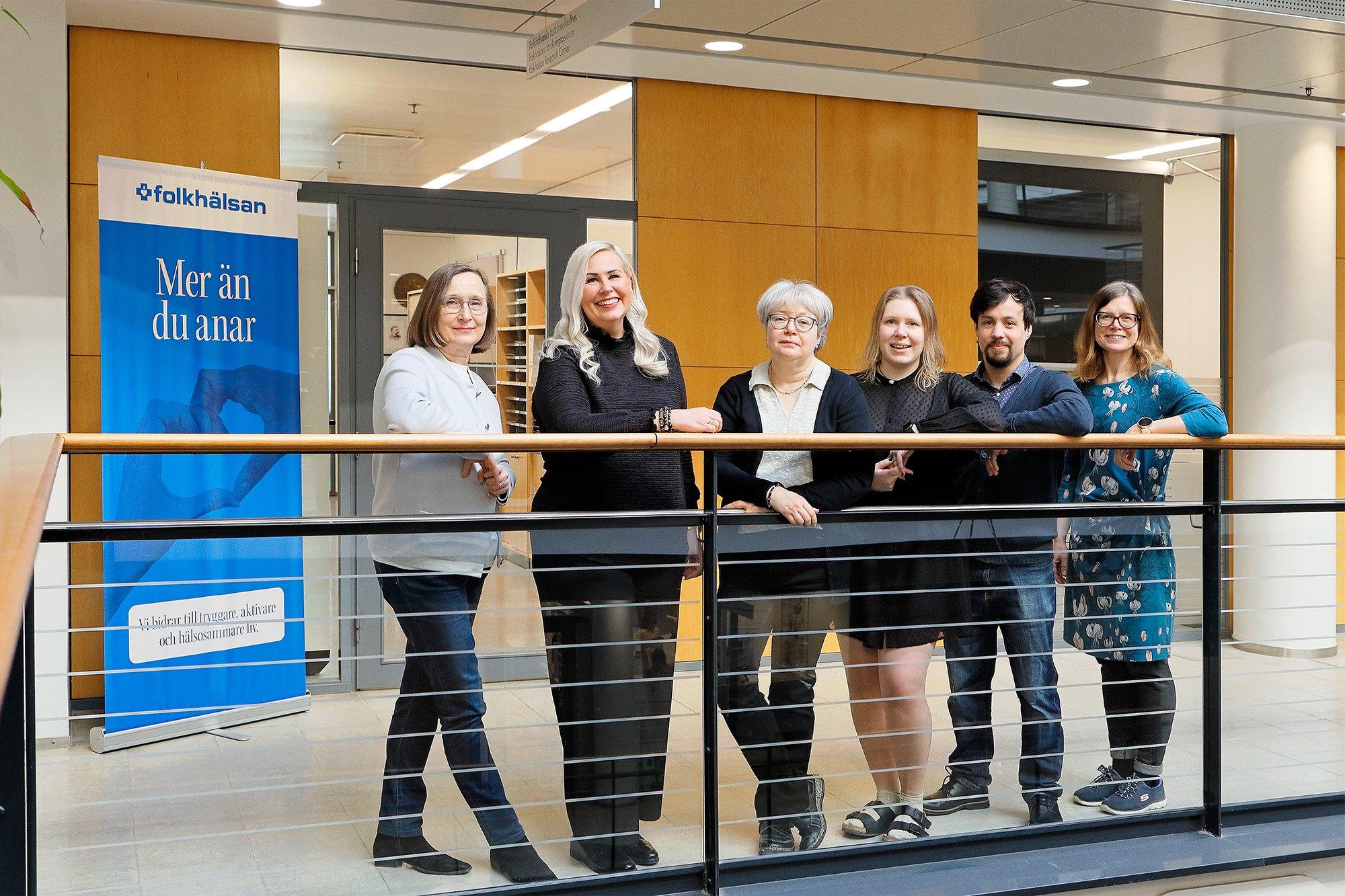 Six people are standing side by side in front of a railing in a bright hallway at Folkhälsan, with a banner in the background that reads “Mer än du anar.” They are smiling and dressed in smart-casual attire, creating a warm and professional group atmosphere